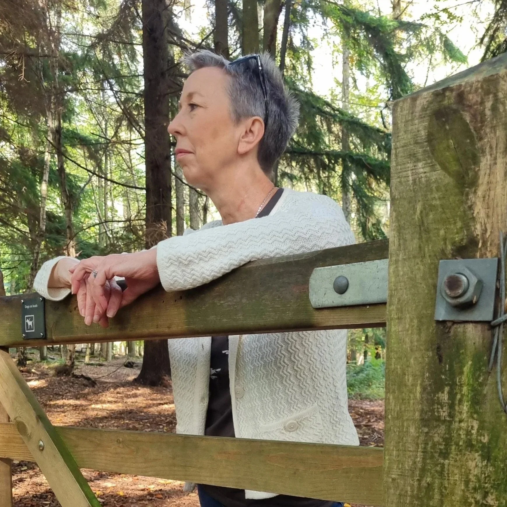A woman with short gray hair and glasses on her head leaning on a wooden railing in a forested area.