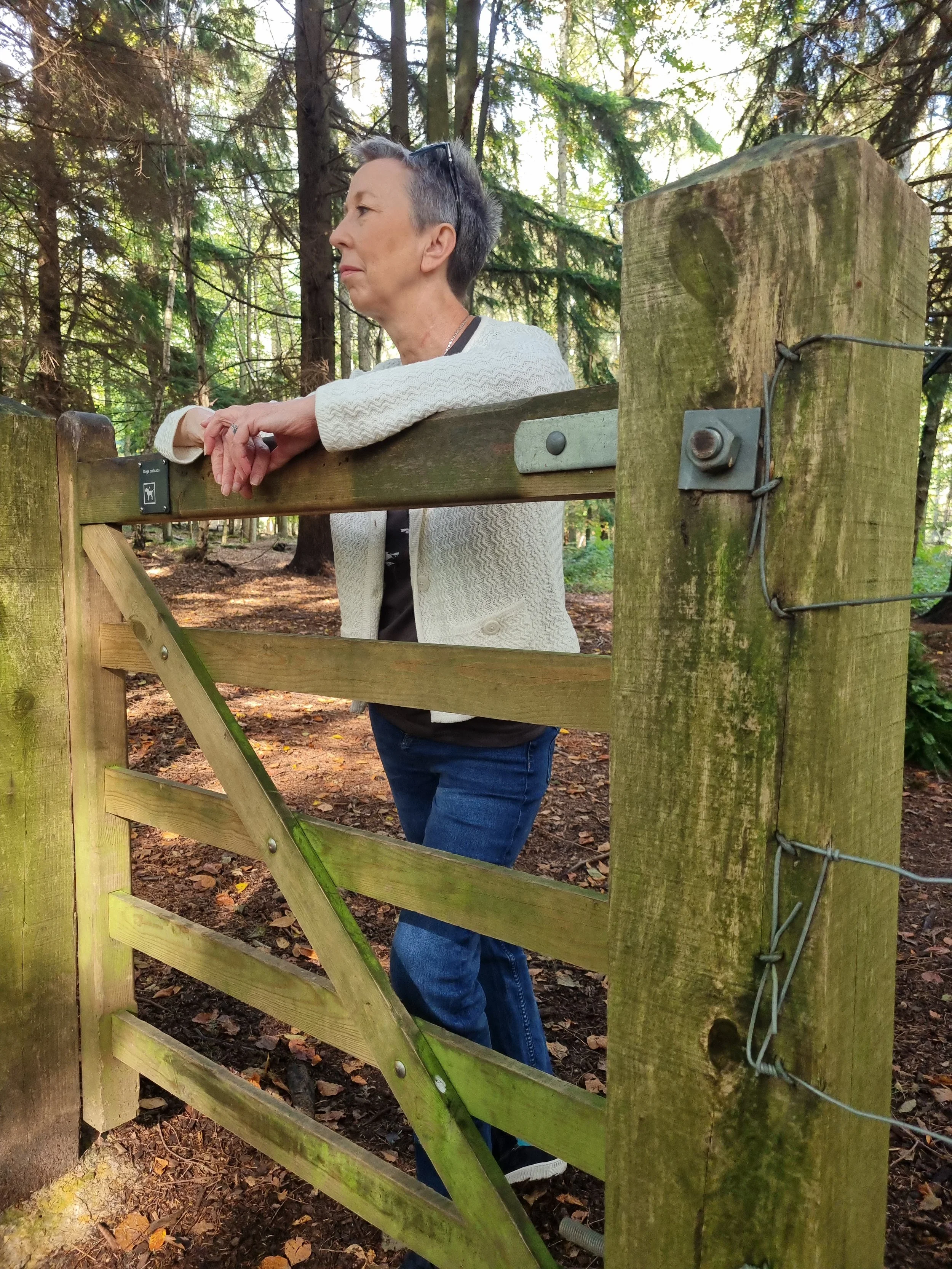 A woman with short gray hair leaning on a wooden gate in a forest, wearing a white sweater and blue jeans.