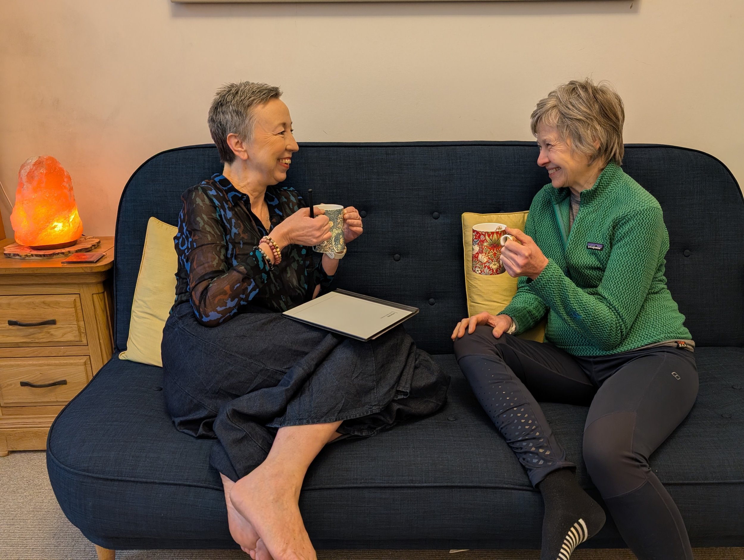 Two women are sitting on a corner sofa, smiling at each other and holding mugs. A salt lamp is on a side table to their left.