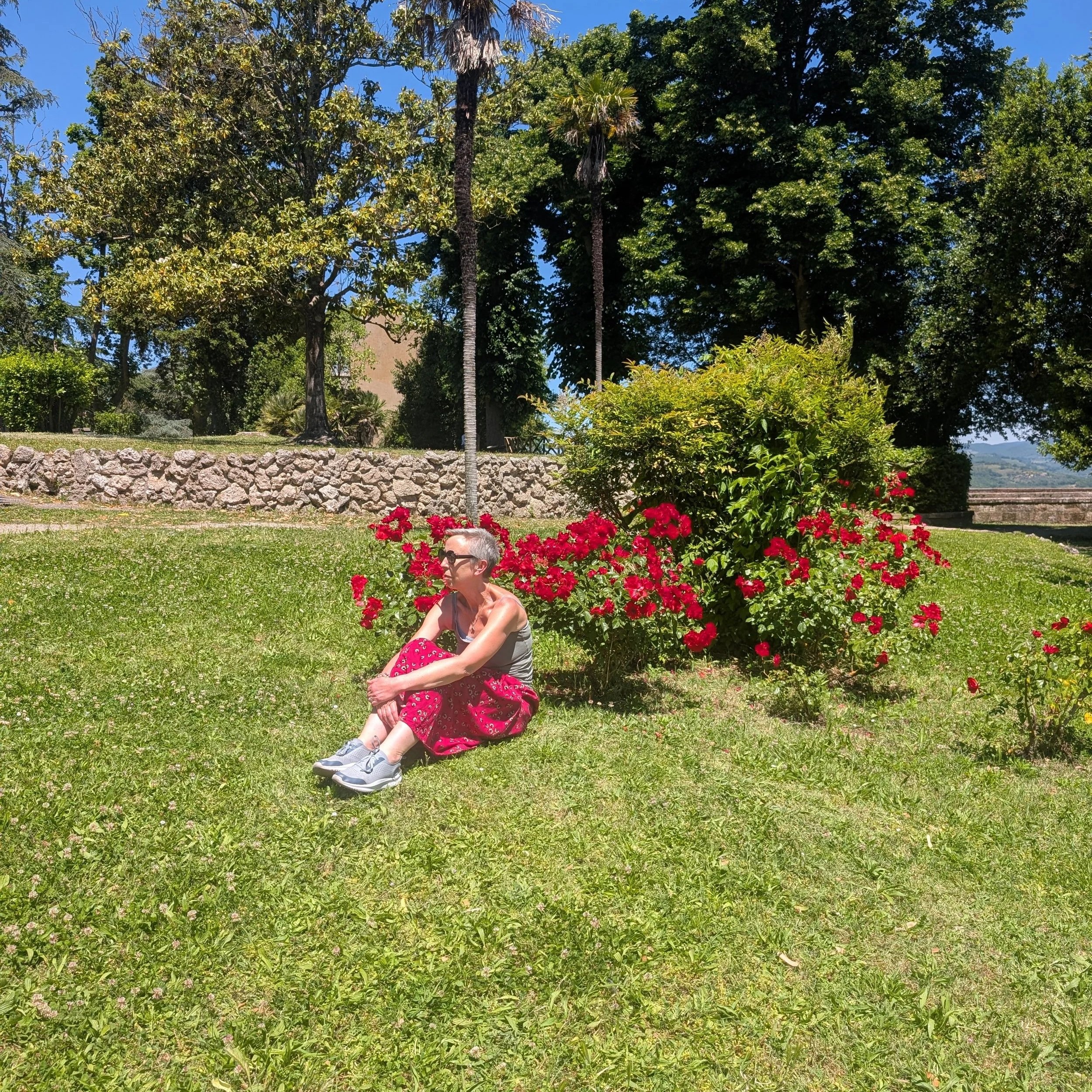A woman sitting on grass with red flowers nearby, surrounded by trees under a clear blue sky.