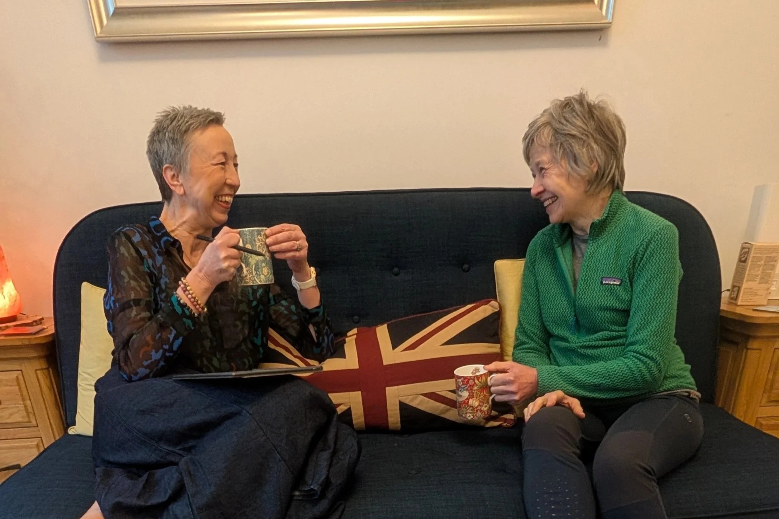 Two women sitting on a blue sofa, laughing and talking to each other. One woman is holding a mug, and the other has a Starbucks cup. They are in a cozy room with a painting and a lamp in the background.