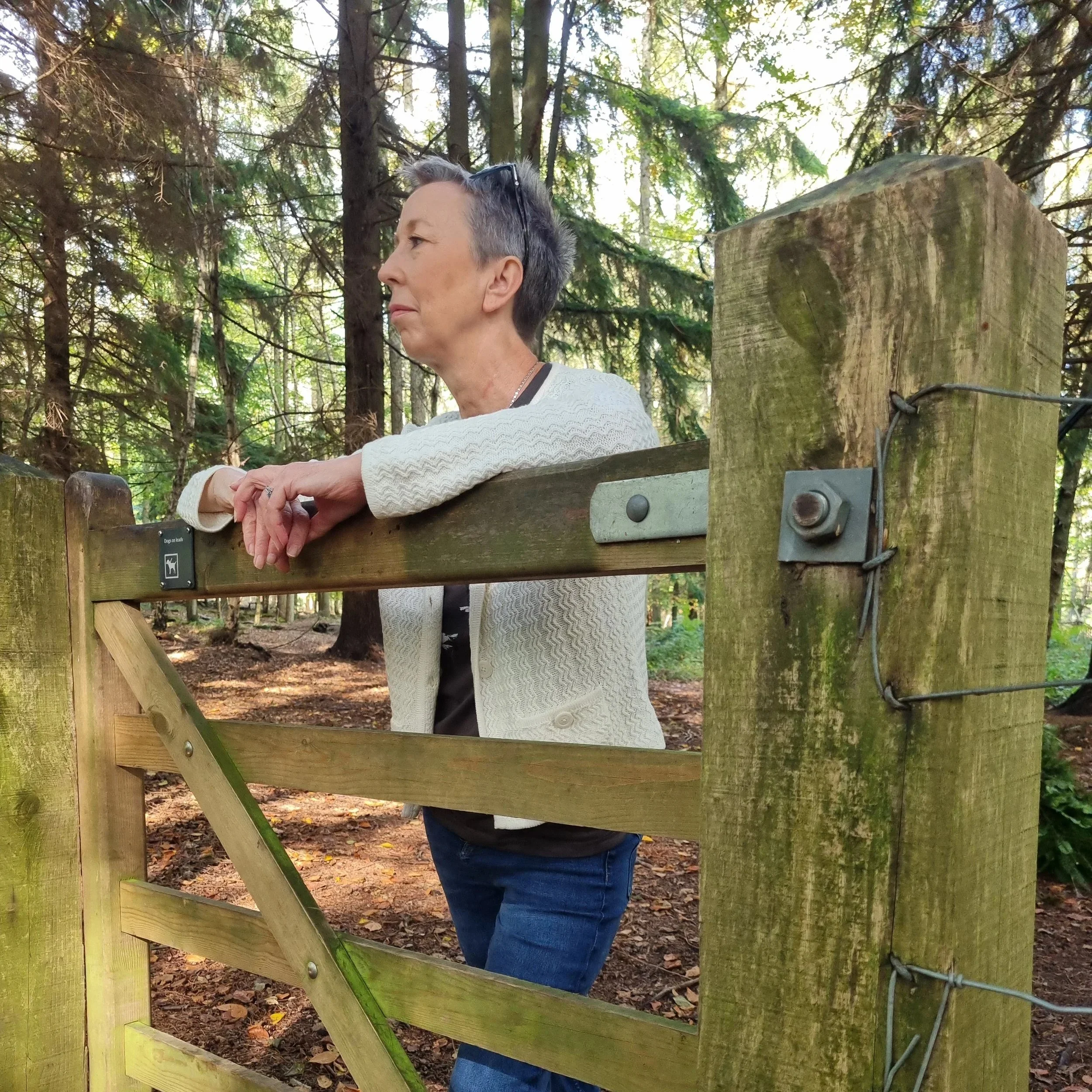 A woman with short gray hair, glasses on her head, wearing a white sweater and blue jeans, leaning on a wooden fence in a forested area with tall trees and green leaves.
