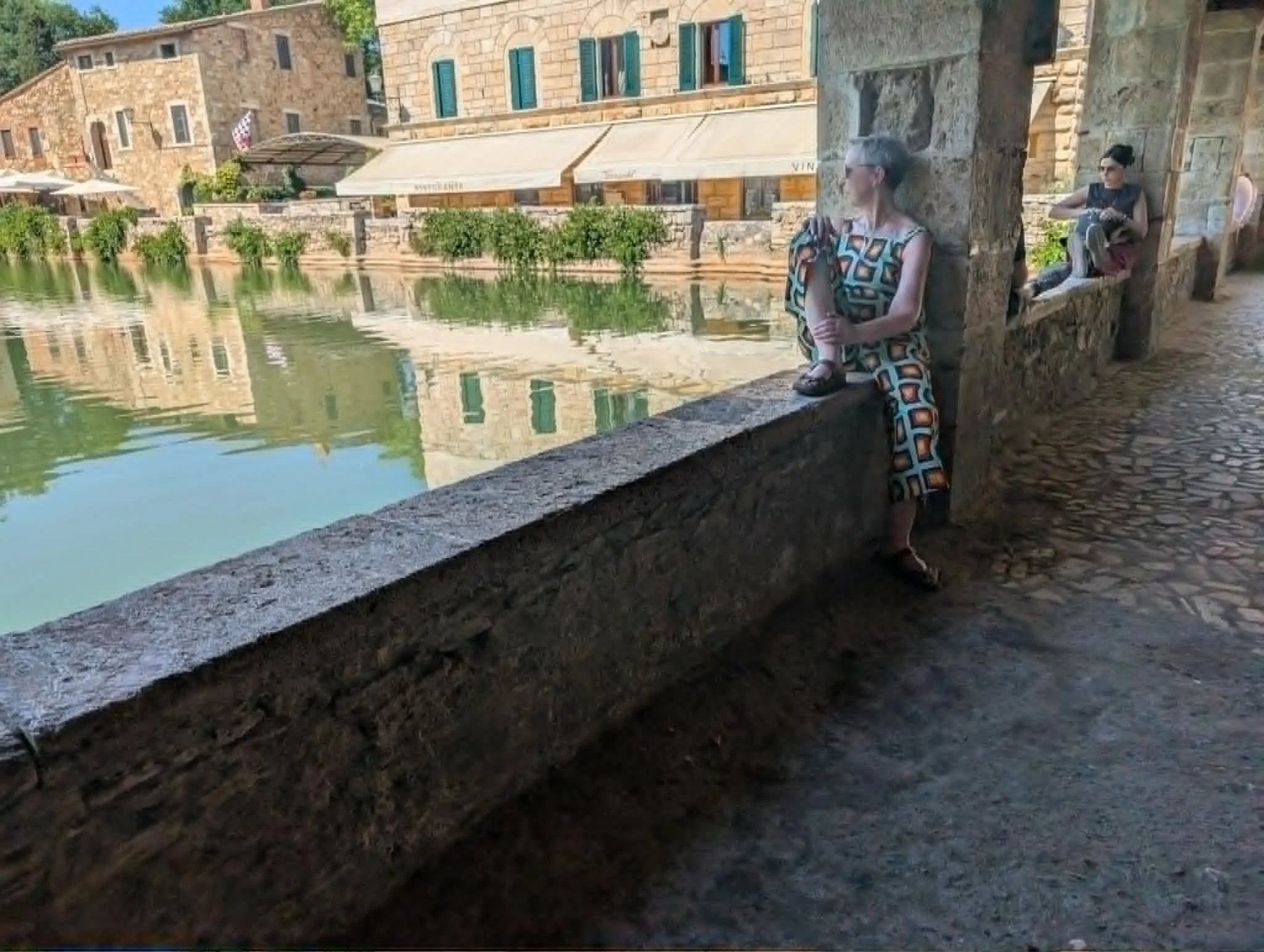 Two women sitting on a stone ledge by a waterway in an old town with stone buildings and green shutters.