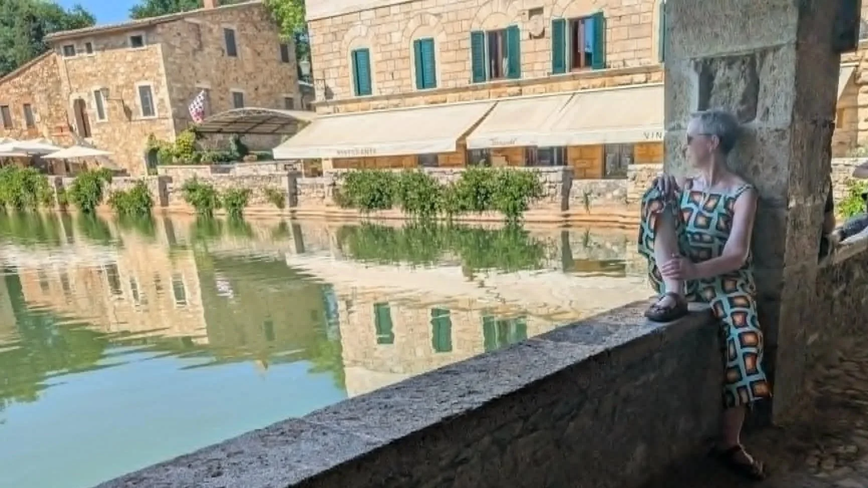 A woman with short gray hair sitting on a stone ledge beside a calm river, wearing sunglasses and a patterned dress. Behind her are old stone buildings with green shutters and a small outdoor café with umbrellas.