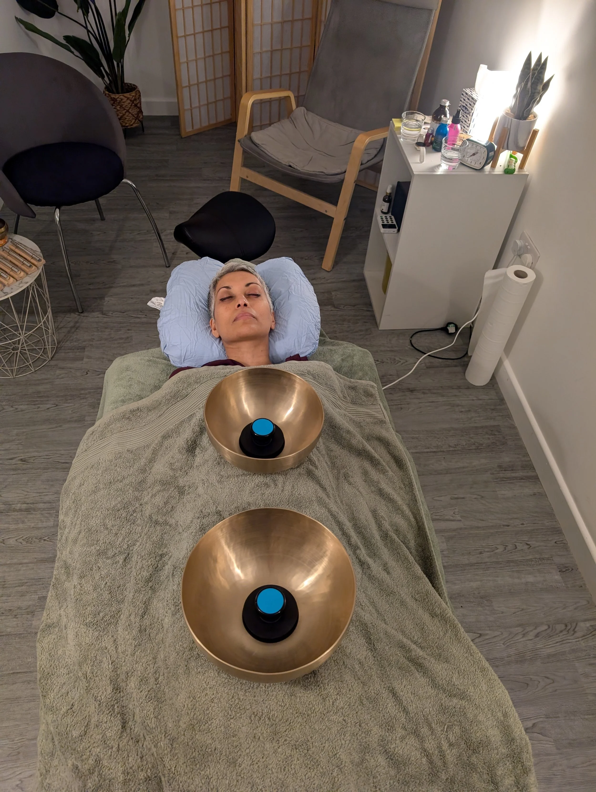 A woman with gray hair is lying on a massage table with her eyes closed, receiving sound therapy with two large singing bowls placed on her stomach. The room has a gray wood floor, a green towel on the table, and a white shelf with various bottles, a clock, and a potted plant. There are chairs, a folding screen, and a tall plant in the background.