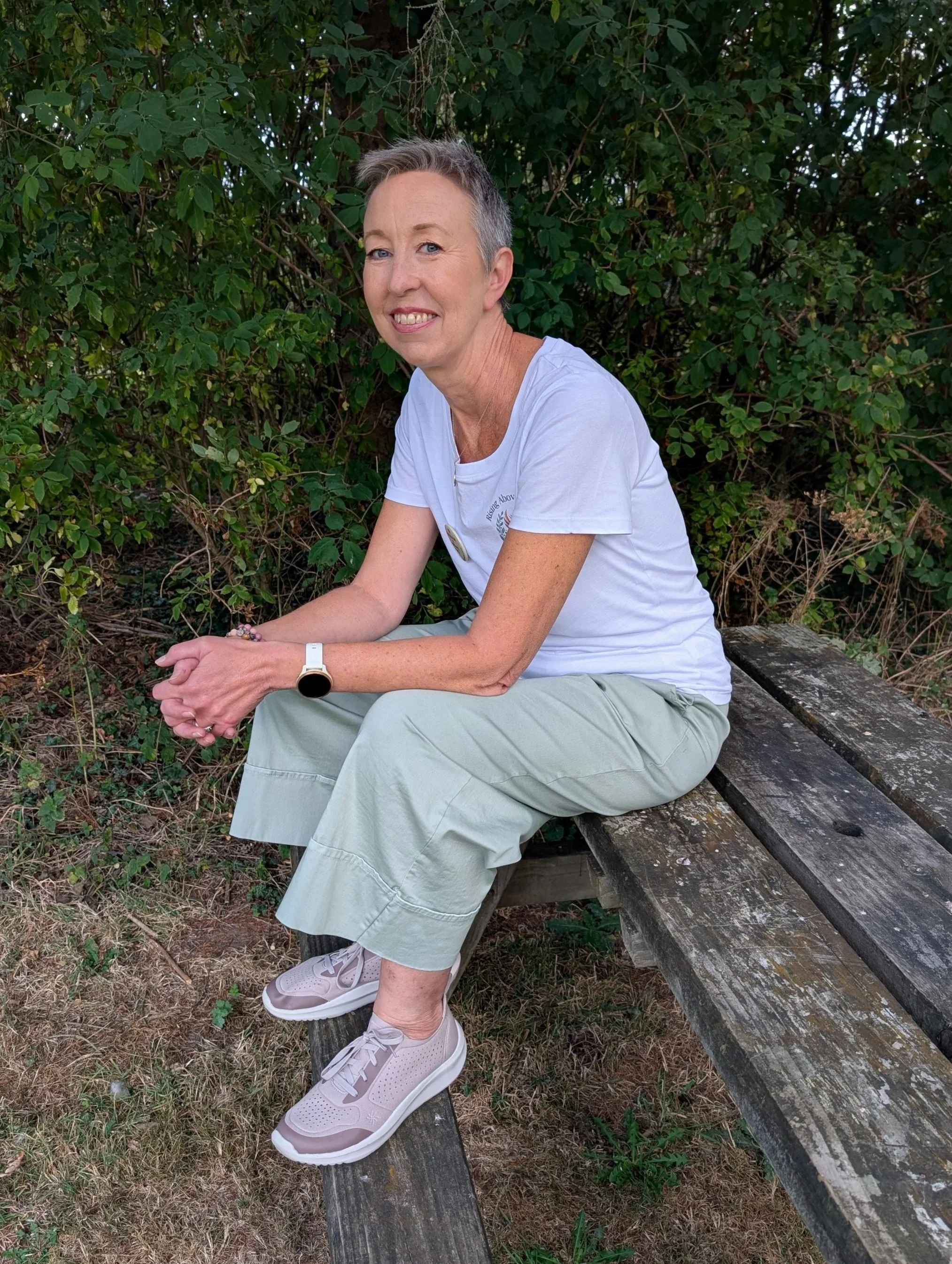 A middle-aged woman with short gray hair, wearing a white t-shirt and light-colored pants, sitting on a weathered wooden bench outdoors with a background of dense green foliage.