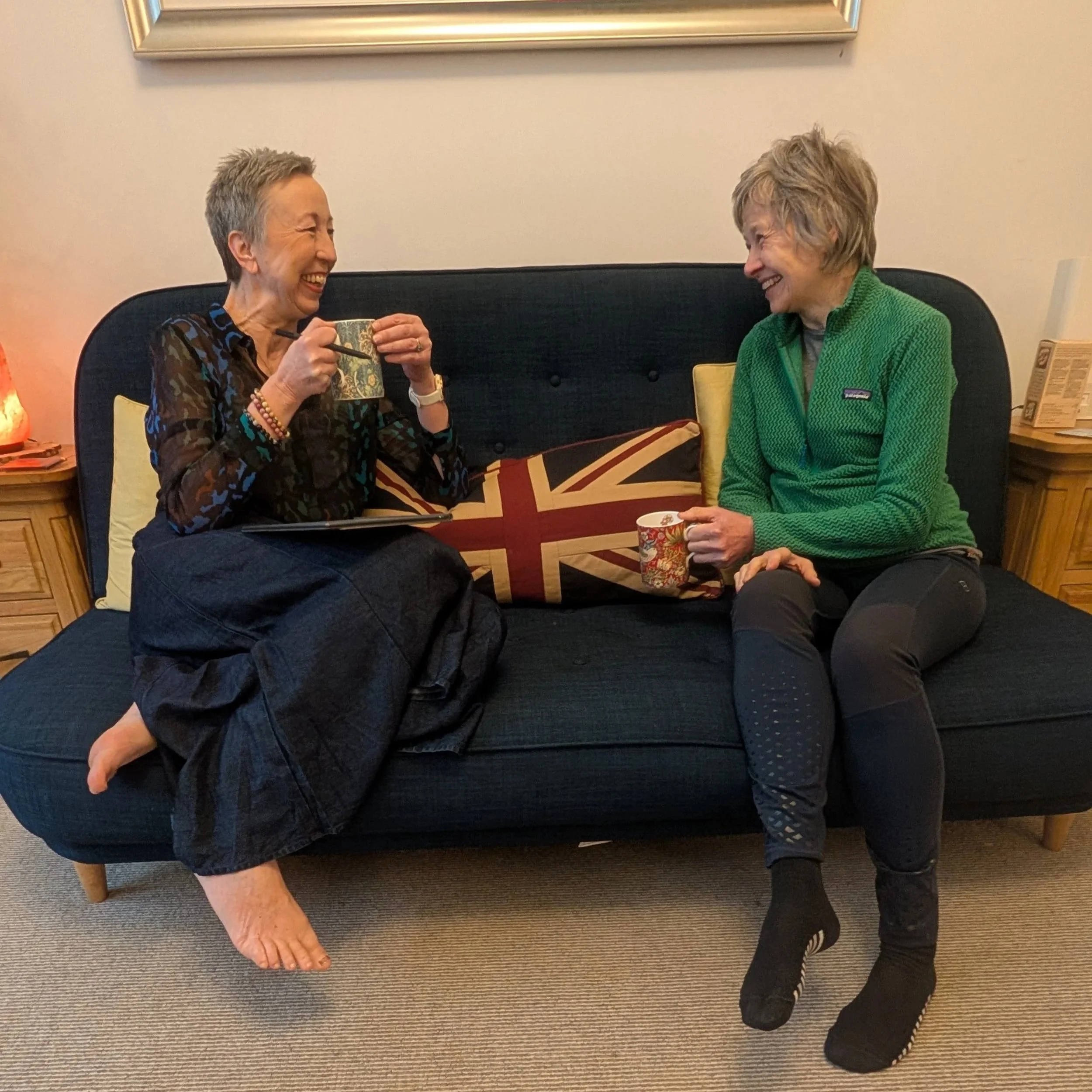 Two women sitting on a black couch, smiling at each other, holding coffee mugs. One woman is barefoot, wearing a dark patterned top and black pants. The other woman is wearing a bright green Patagonia fleece and black pants, with sneakers. There is a pillow with the Union Jack design on the couch. A framed mirror hangs on the wall behind them, and a wooden side table with a salt lamp and a decorative block is visible.