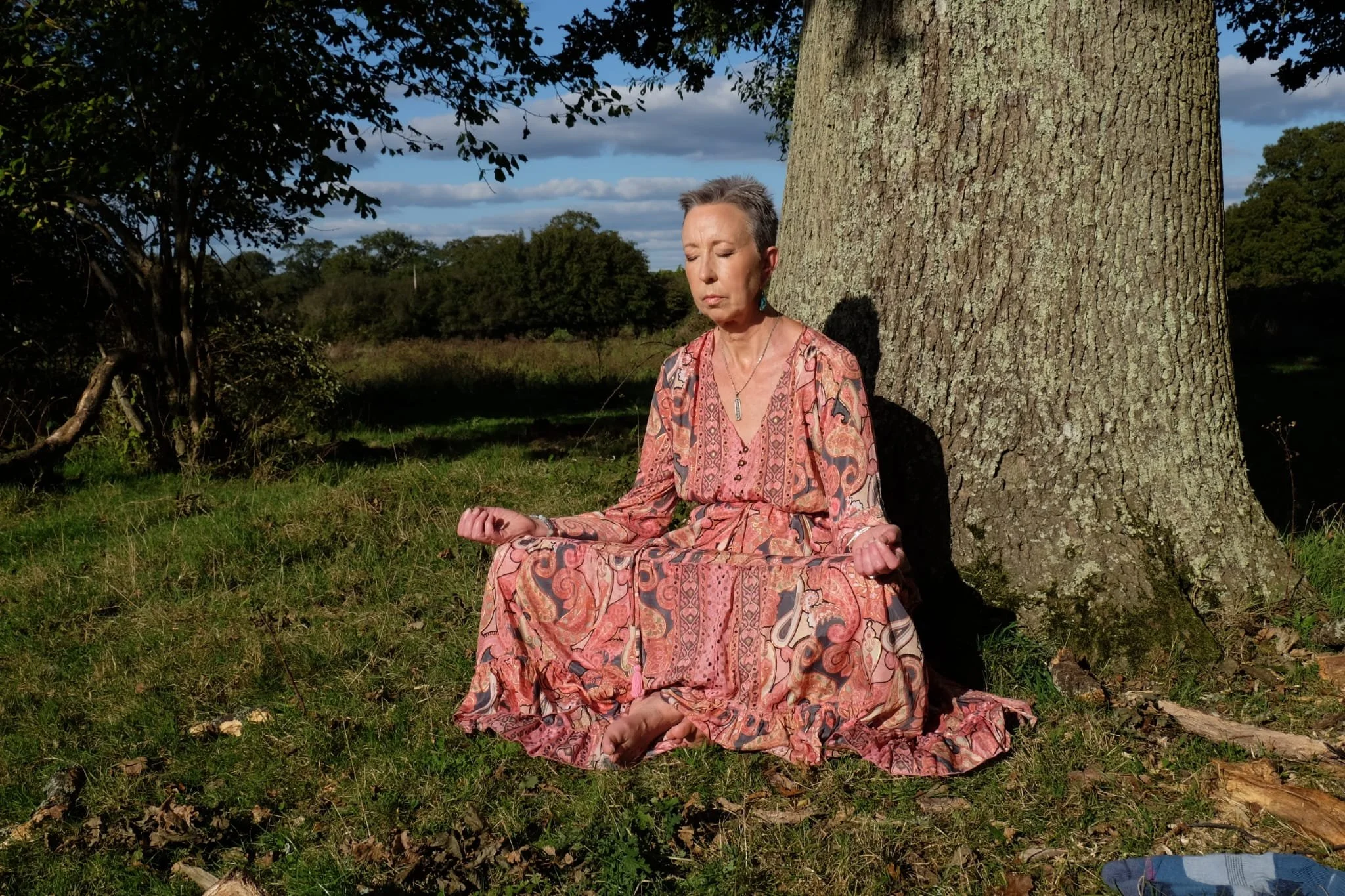 A woman with short gray hair is sitting cross-legged on the grass, meditating outdoors next to a large tree on a sunny day with blue sky and clouds in the background.