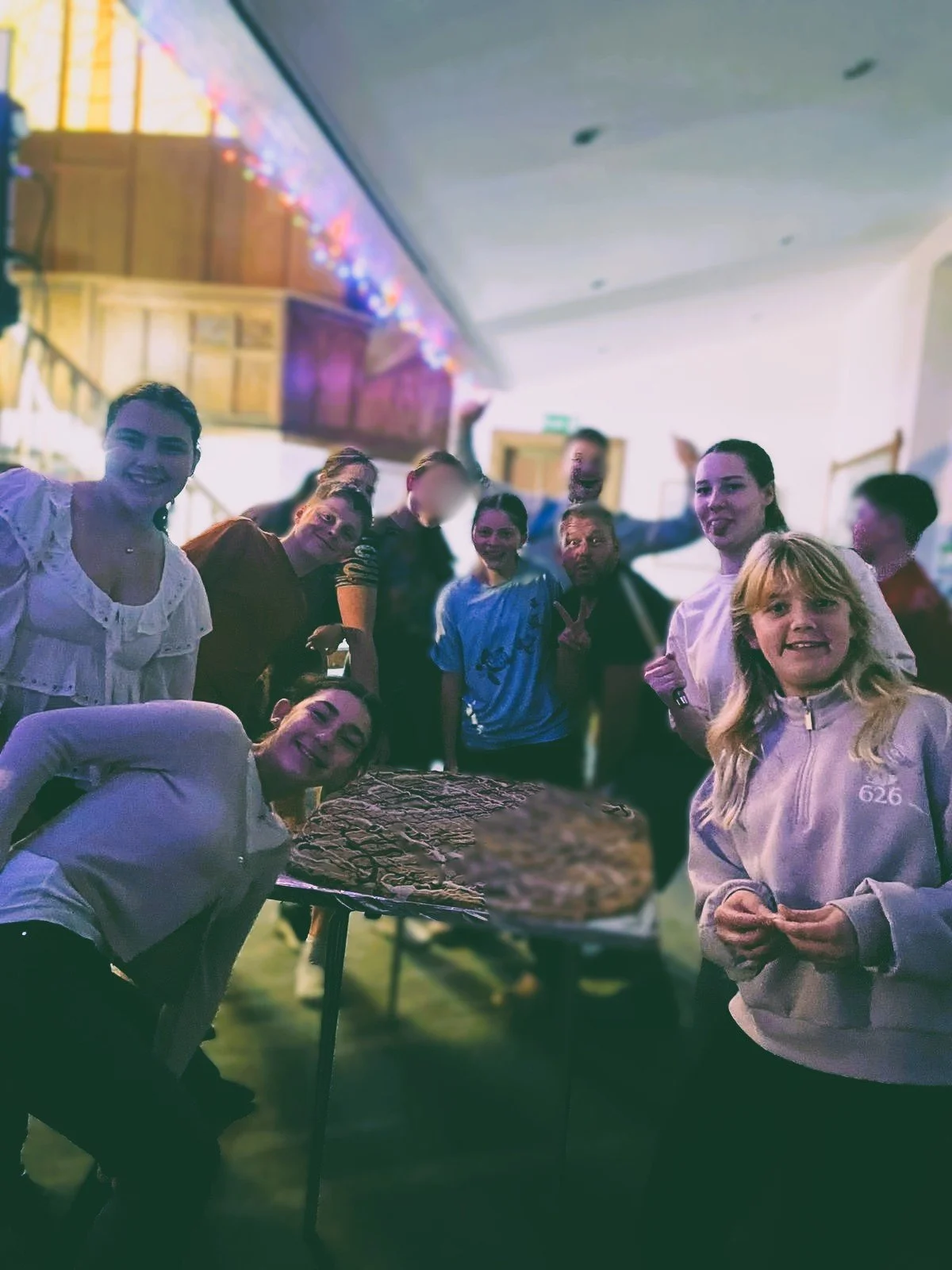 Group of people celebrating with pizza in an indoor setting decorated with string lights.