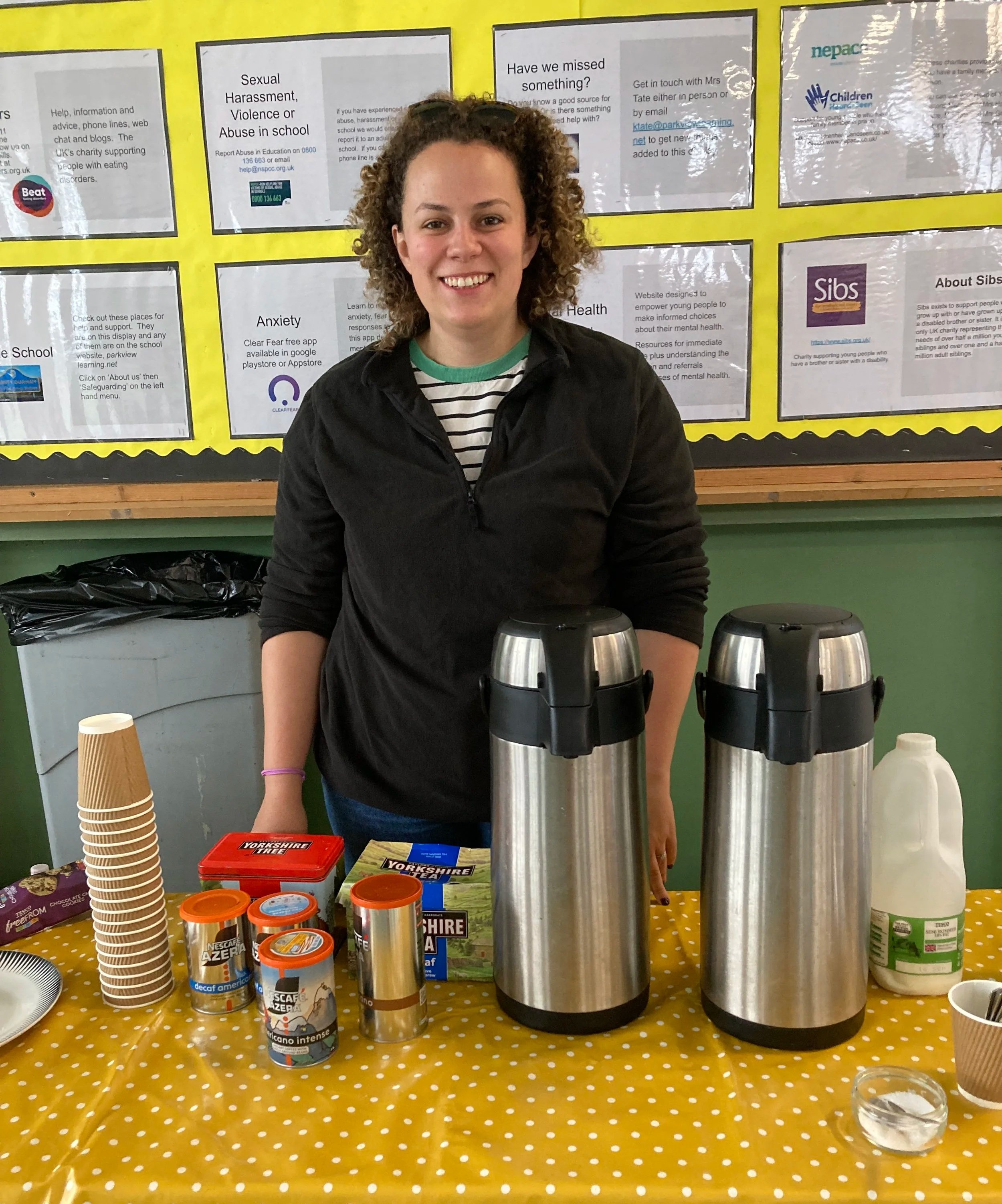 Four people standing in a kitchen with drinks and coffee cups on a counter in the foreground. Two men and two women, all wearing name badges and smiling at the camera. There are coffee pots and pitchers of juice on the counter.