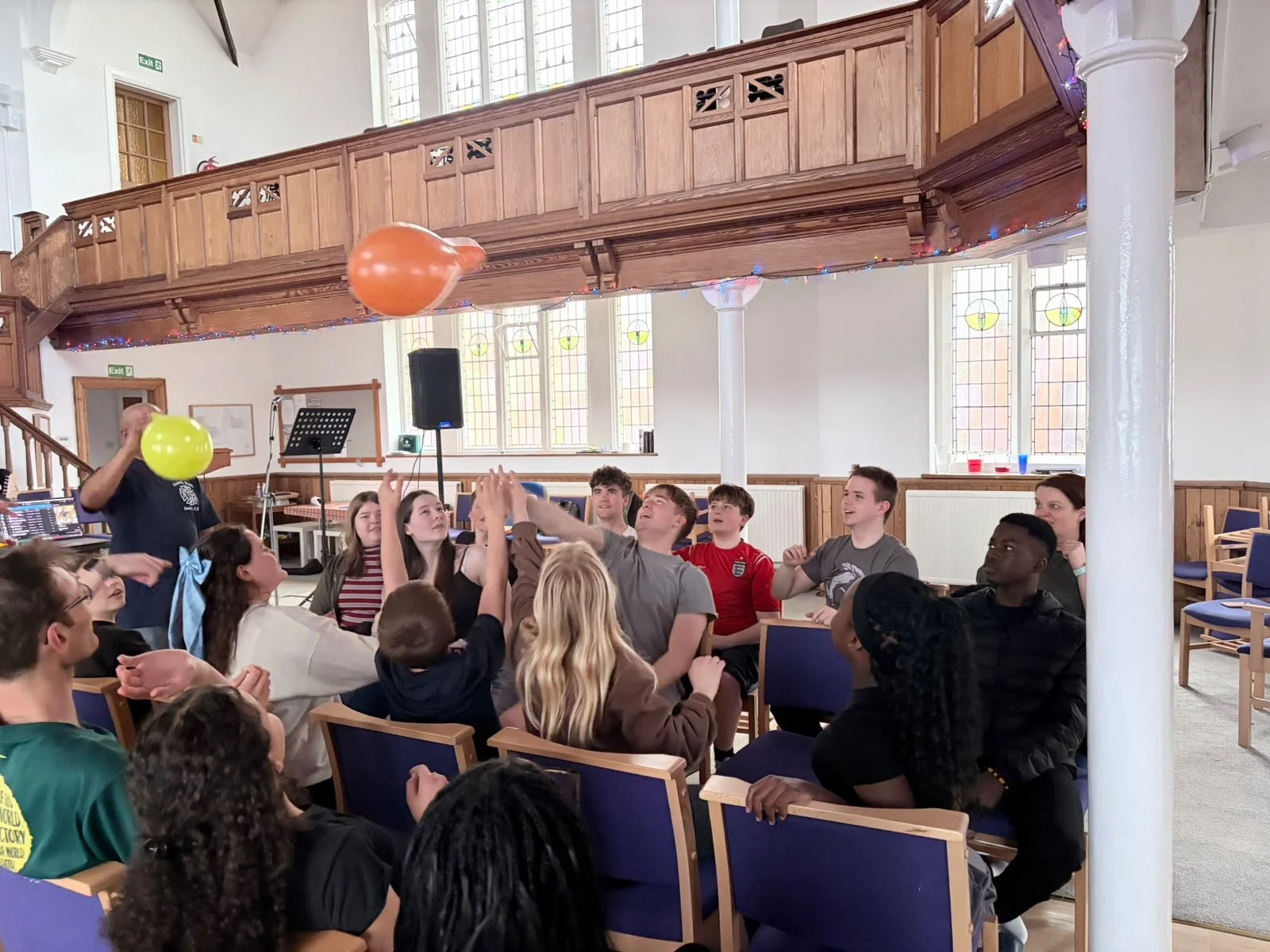 Group of young people sitting in a church or community hall, engaging in a game involving balloons, with smiling and cheering expressions, decorated with string lights and stained glass windows.