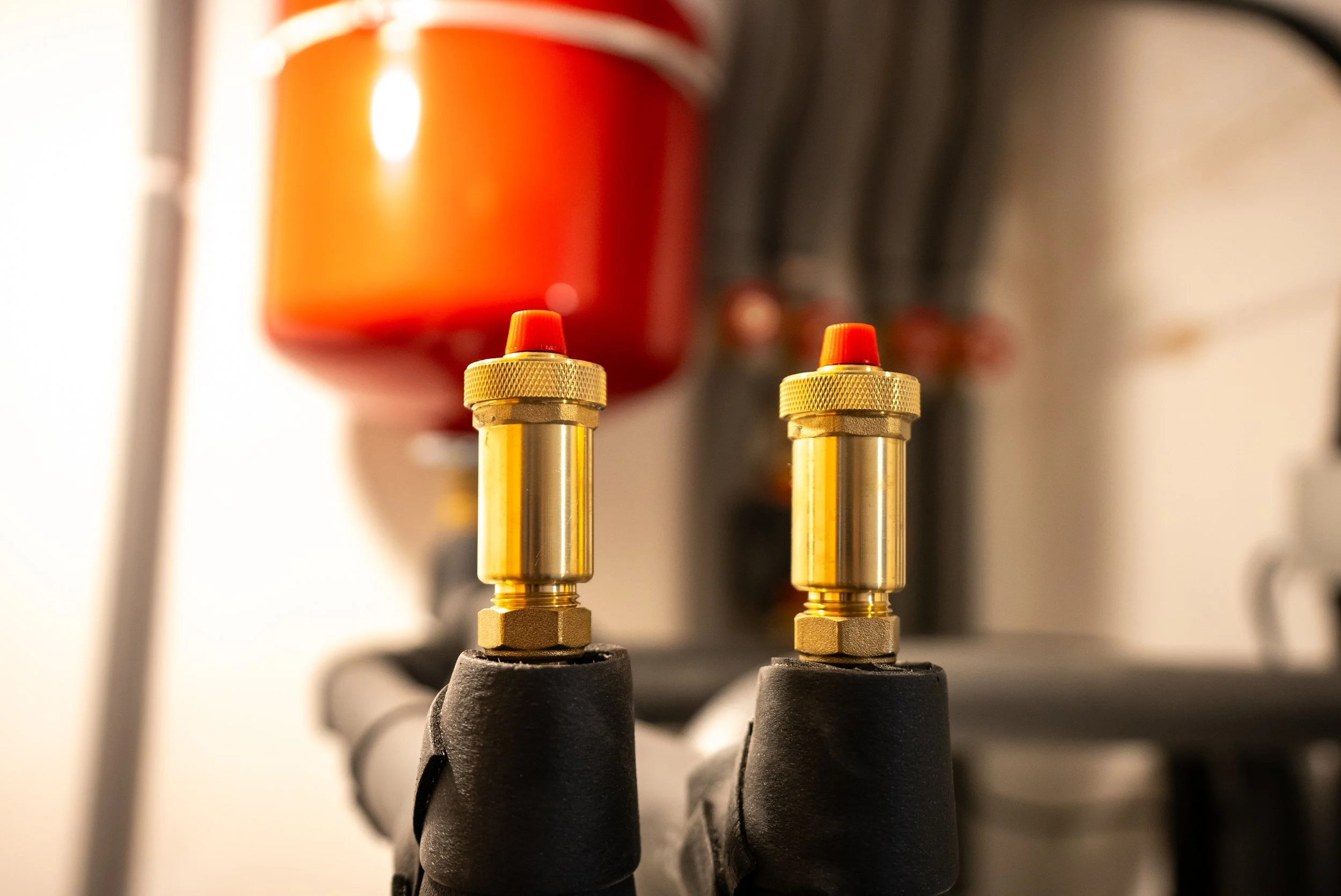 Close-up of two brass valves with orange caps, attached to black handles, with a large red container in the background.
