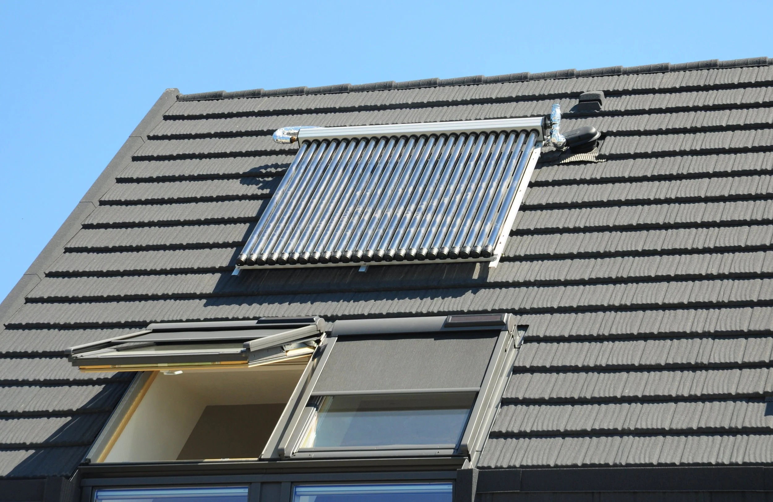 A solar water heater installed on the sloped roof of a building, with a window open below it and a clear blue sky in the background.