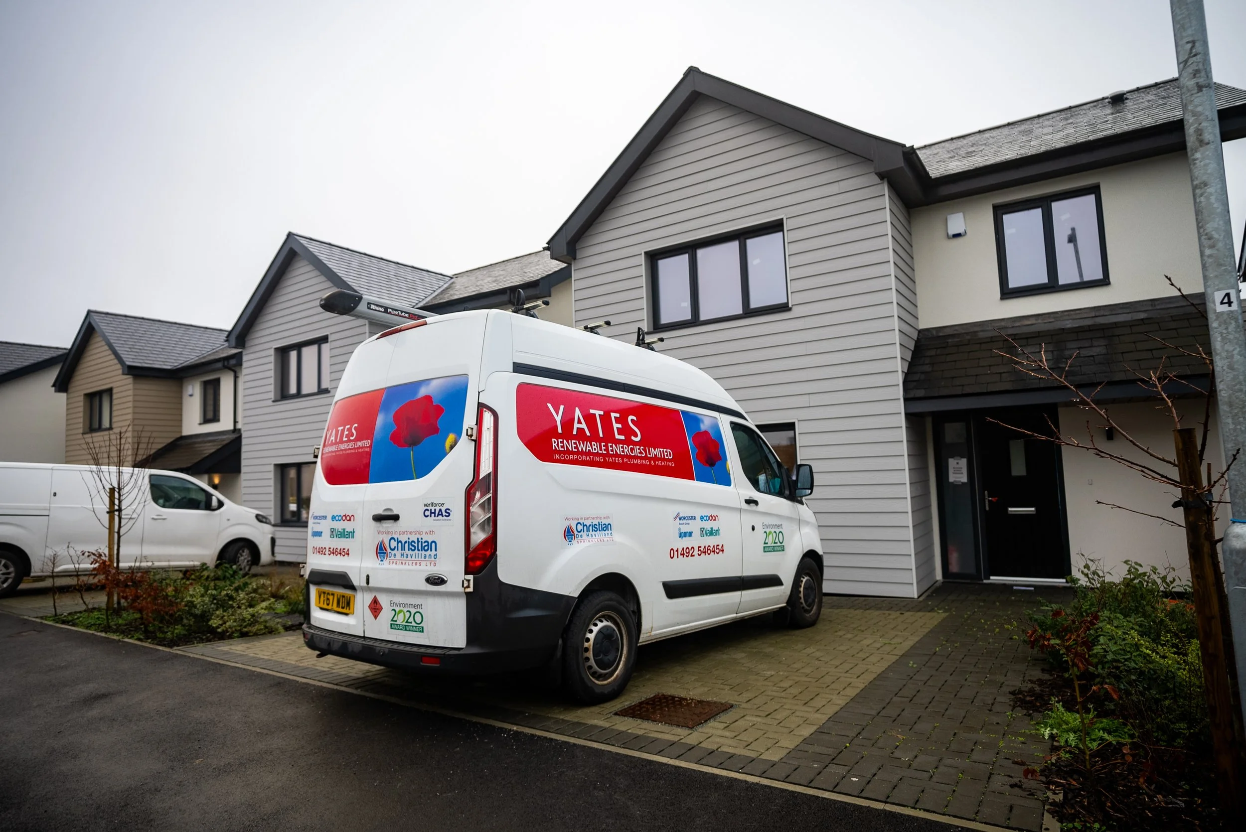 A white van parked in front of a modern residential building with grey and beige siding. The van has branding for Yates Renewable Energies Limited and Christian Haviland Ltd, including logos and contact information.