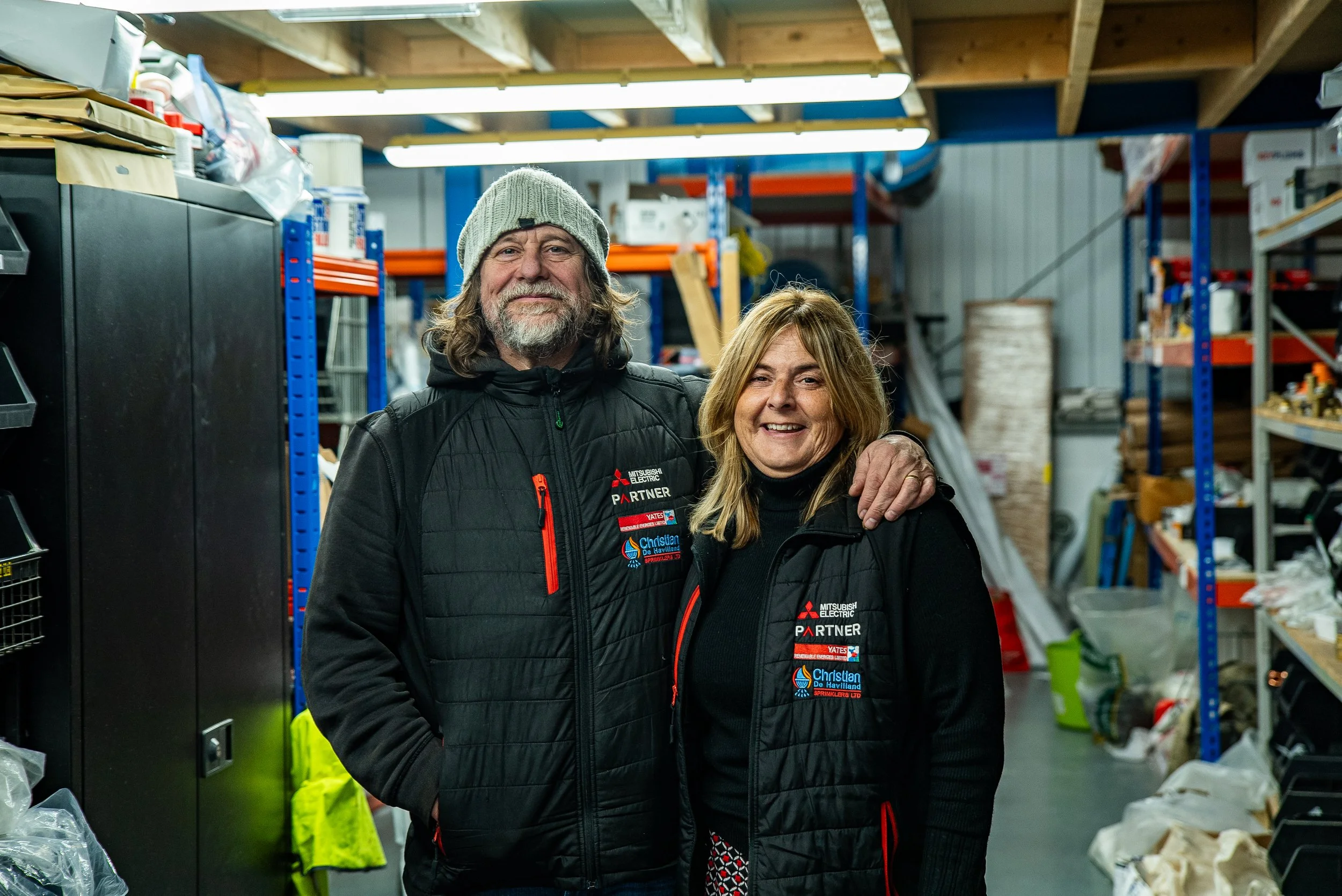 A man and woman standing together in a warehouse, both wearing black jackets with company logos, smiling at the camera.