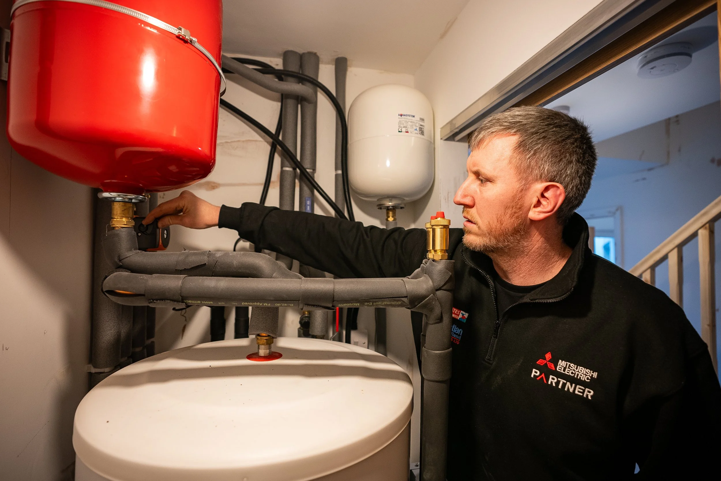 A technician working on a water heater, wearing a black jacket with a Mitsubishi Electric logo, in a residential utility closet.