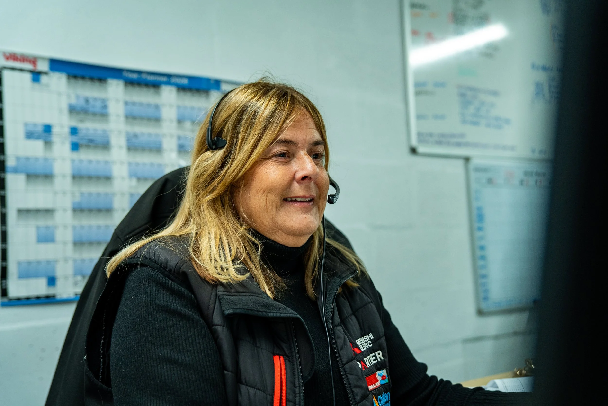Woman wearing a black vest and headset sitting at a desk in an office or control room with whiteboards and charts in the background.