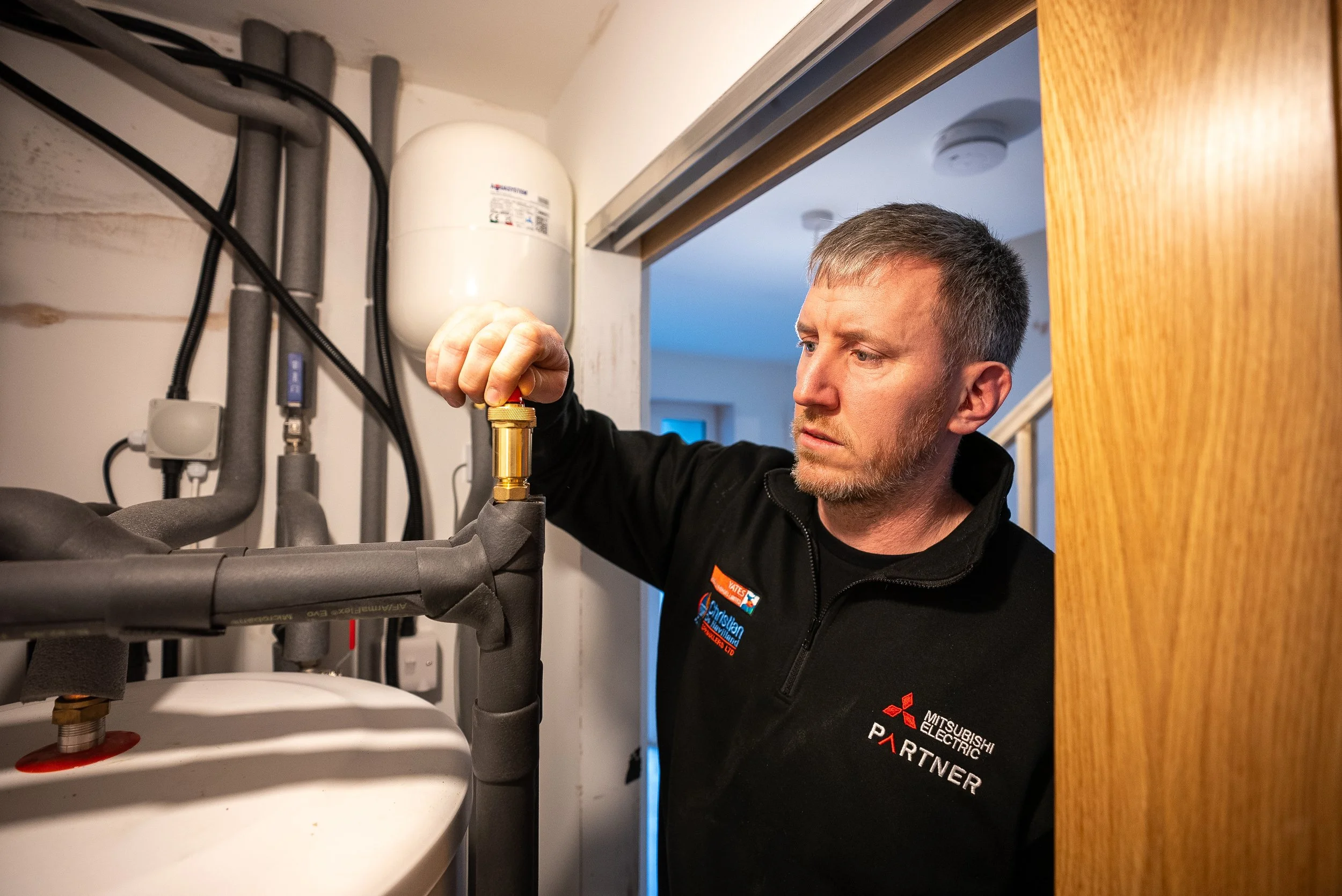 A technician inspecting or repairing plumbing or heating pipes in a utility room wearing a black shirt with Mitsubishi Electric branding.