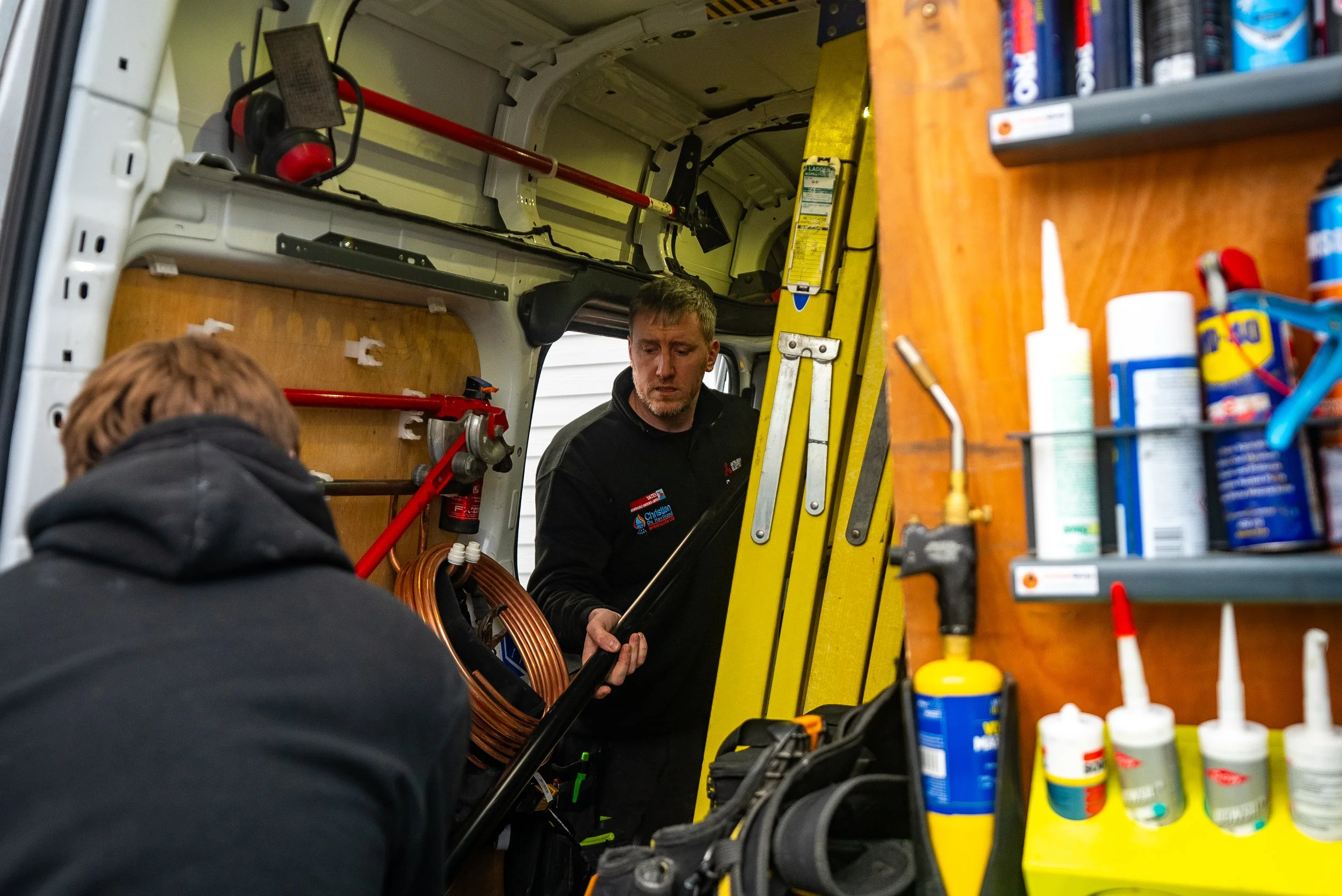 Two men working inside a utility vehicle, one holding a long tool, with various tools and supplies around them.