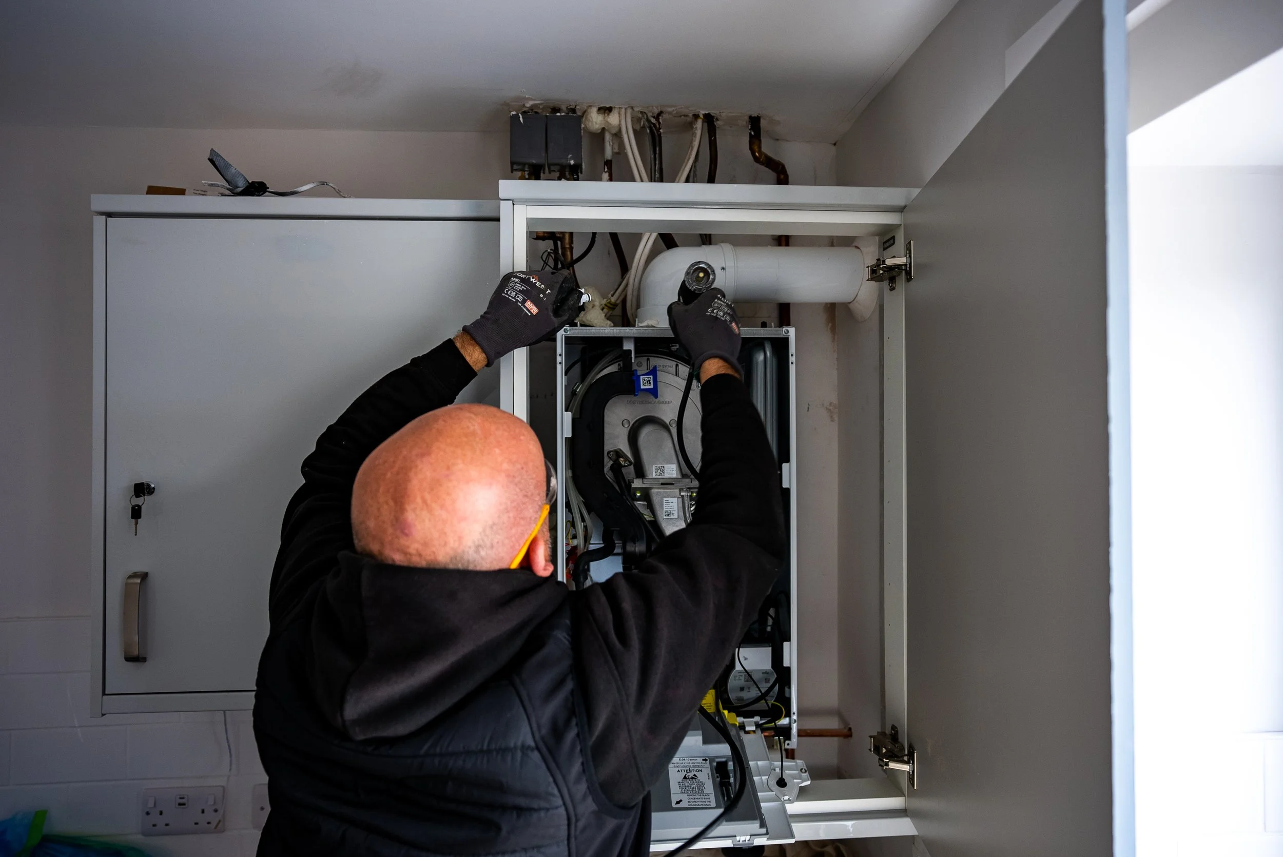 A technician working on a kitchen boiler or furnace, wearing black gloves and a black jacket, using tools inside an open white cabinet.