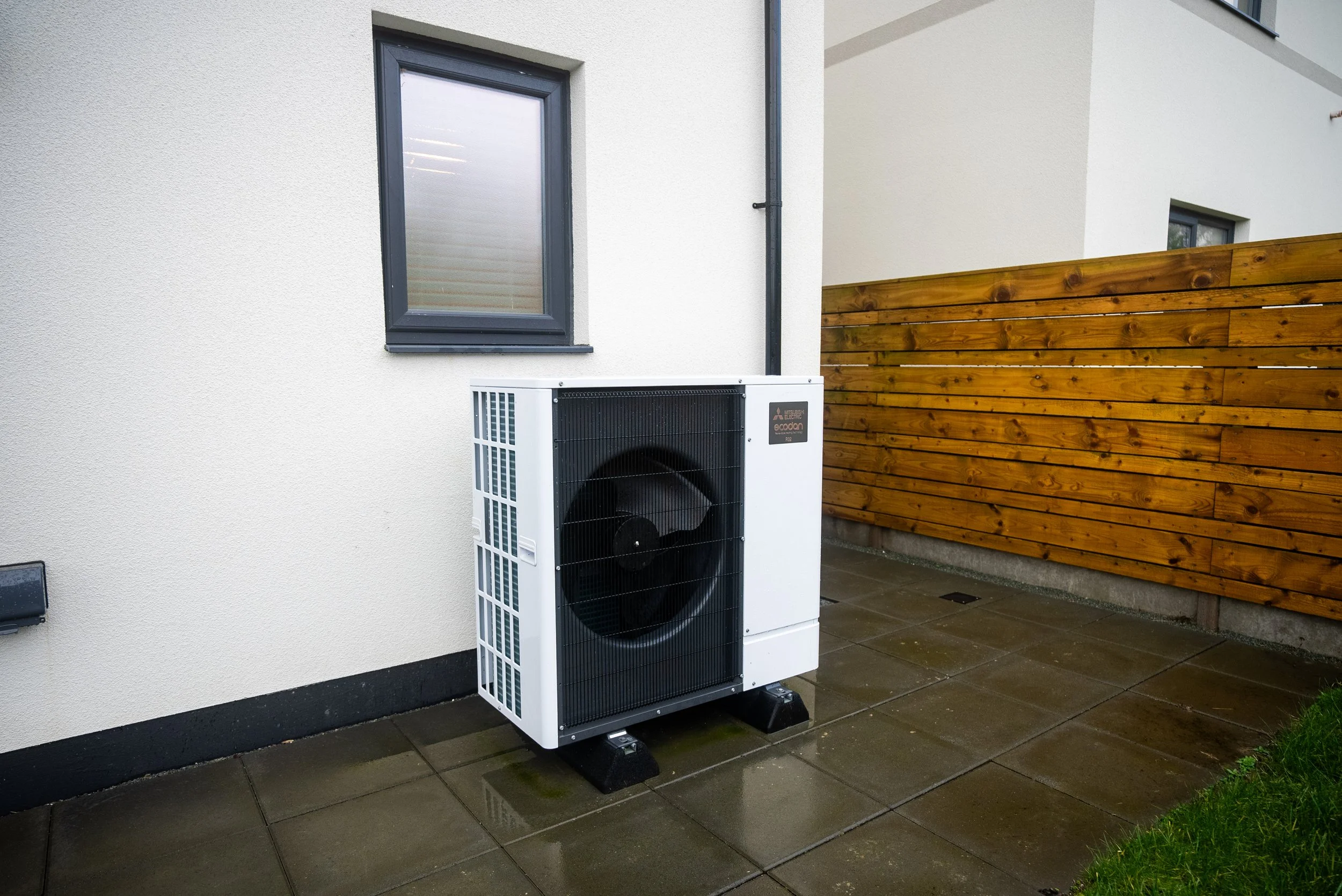 Outside view of an air conditioning unit installed on a patio next to a beige house wall and a wooden fence.