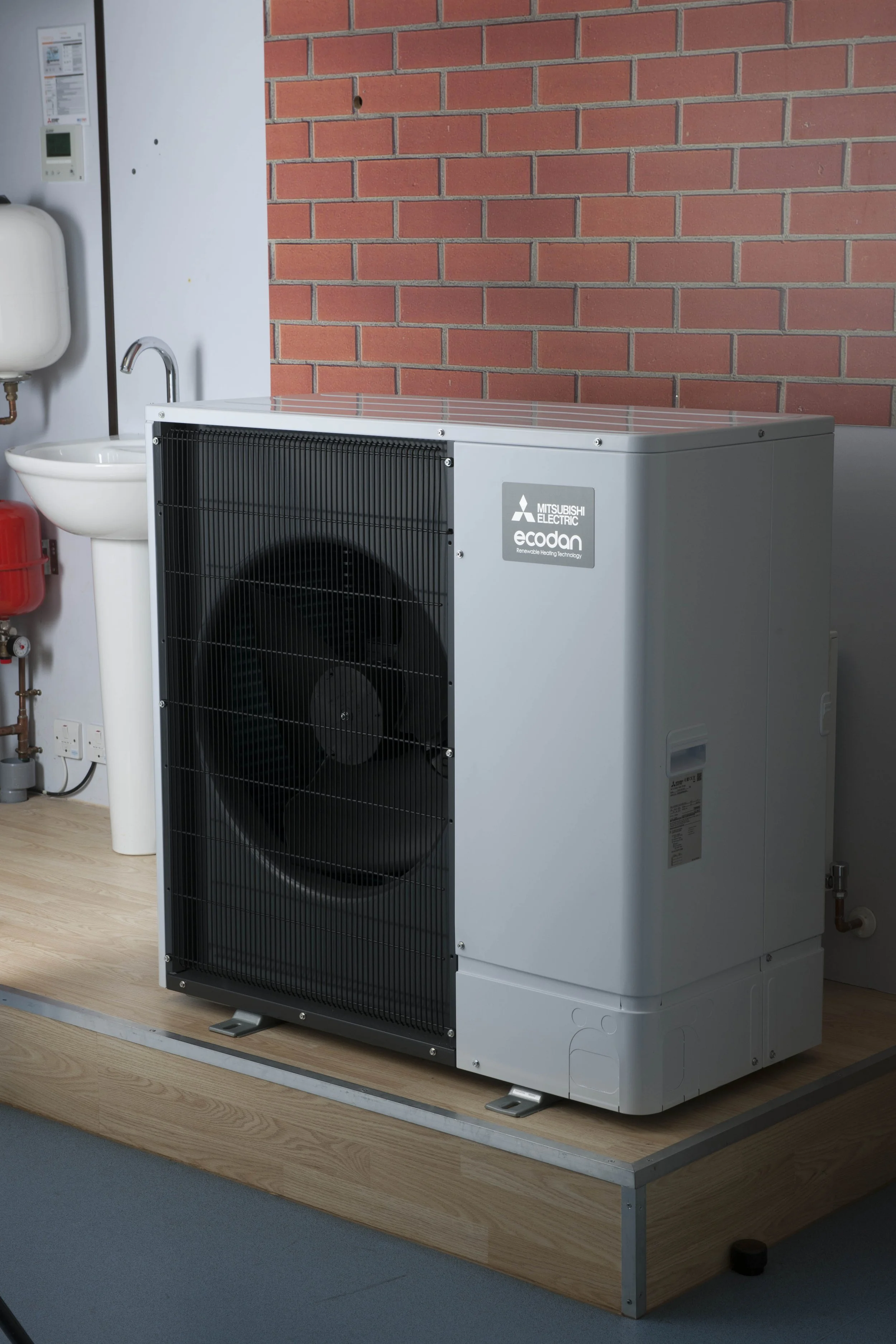 Outdoor Mitsubishi Electric Ecodan heat pump on a wooden platform against a brick wall.