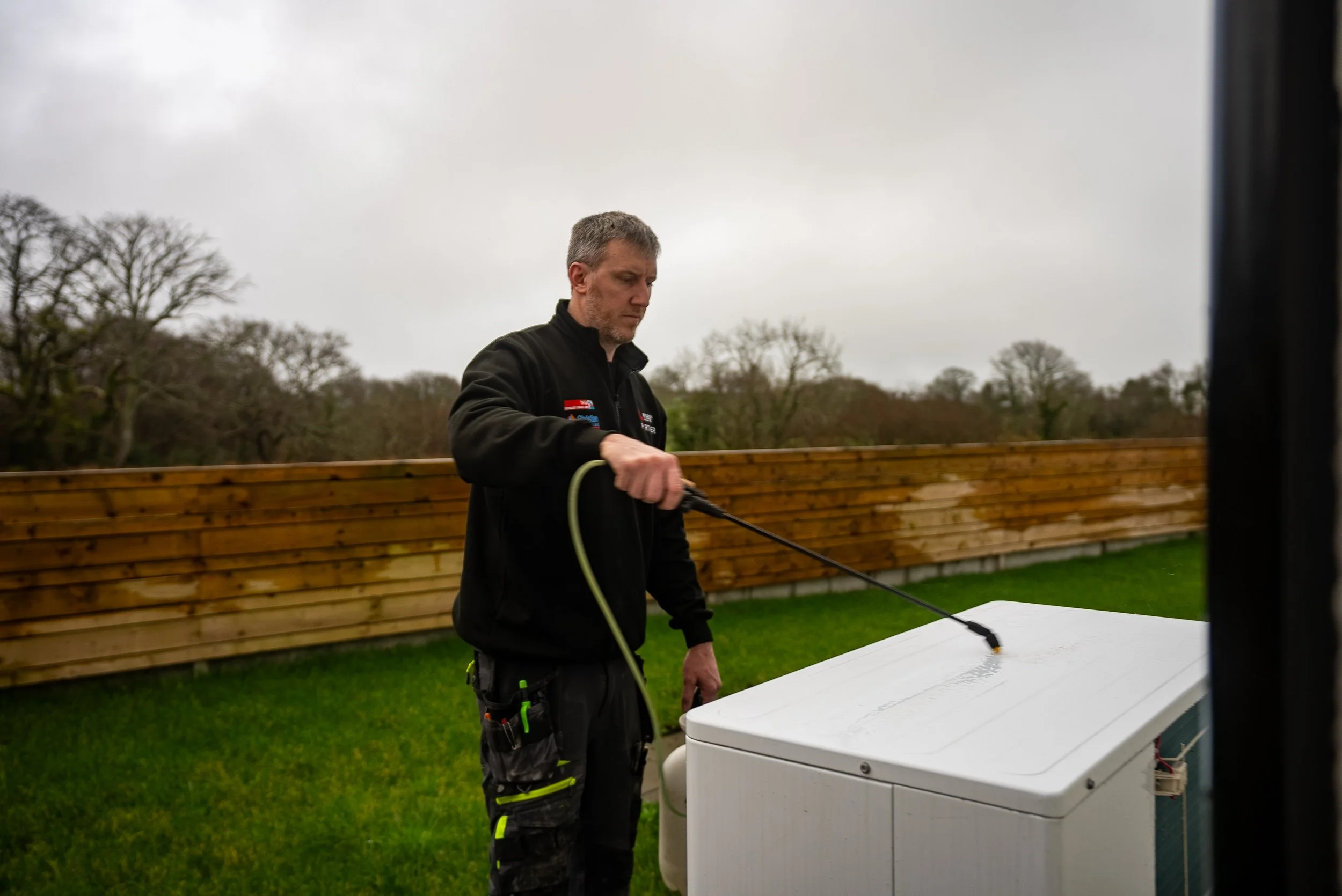 A man cleaning the top of an outdoor air conditioning unit on a lawn, with a wooden fence and trees in the background on a cloudy day.