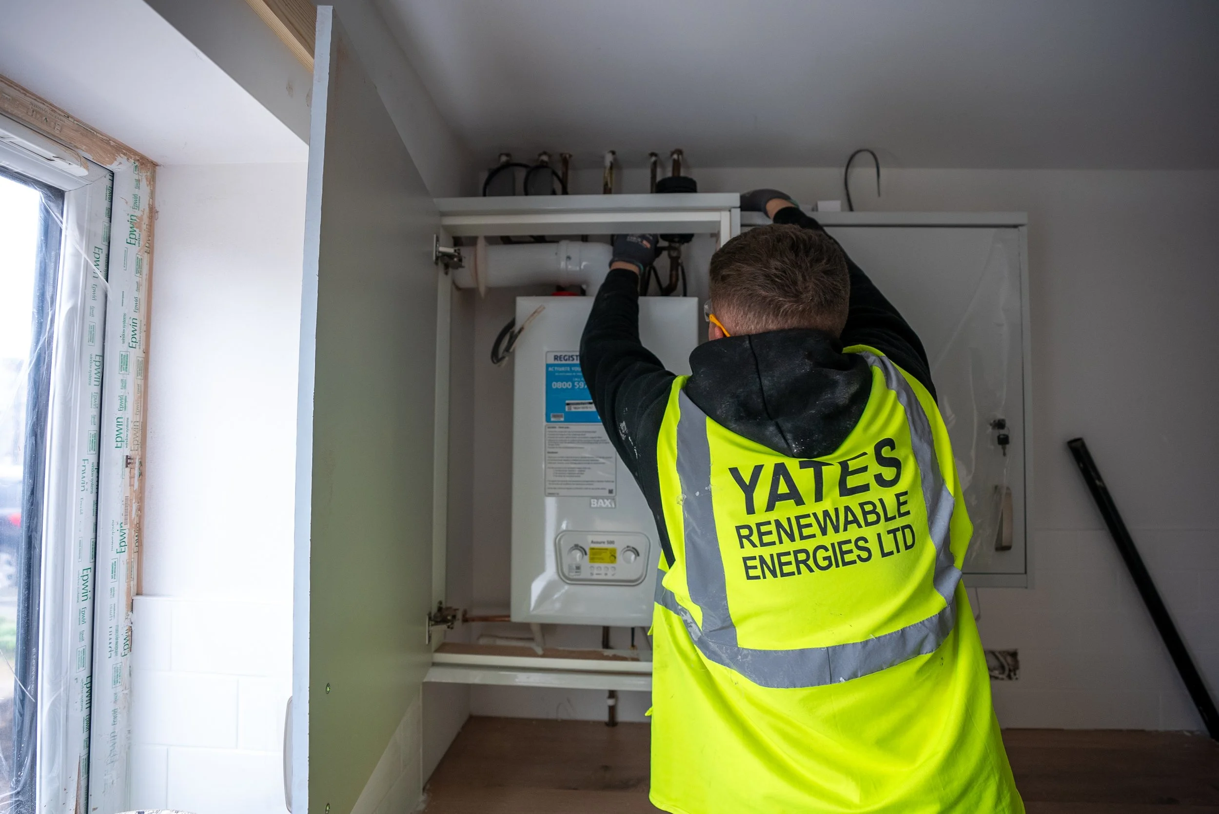 A worker installing or repairing a boiler or heating unit in a room with a window, wearing a high-visibility vest with the company name YATES RENEWABLE ENERGIES LTD on the back.