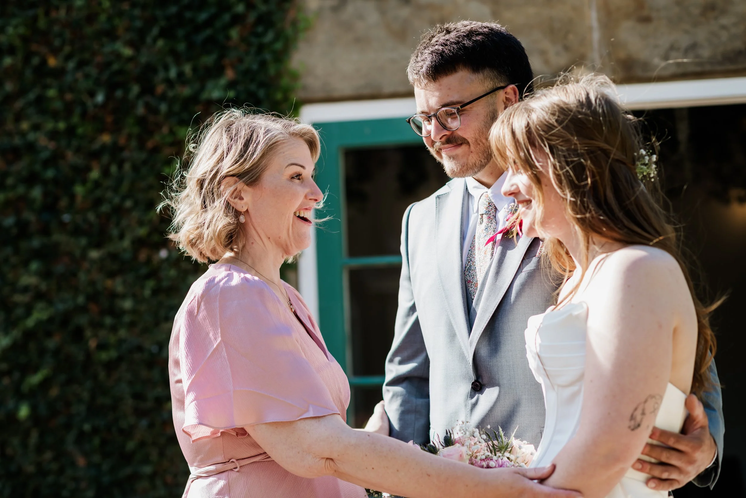 Tammy is smiling and in conversation with the bride and groom soon after their wedding , outdoors with a green hedge and building in the background.
