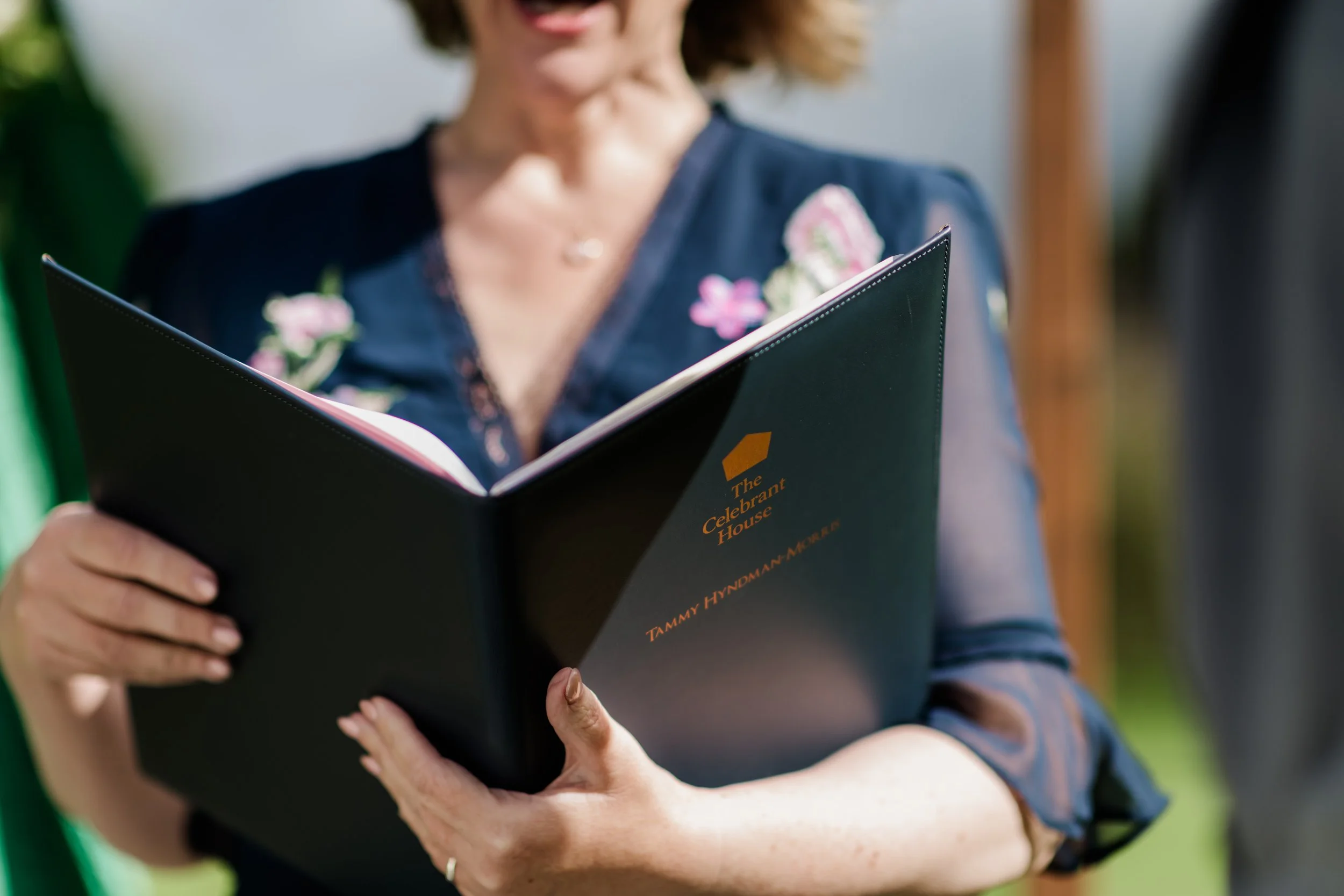 Close-up of Tammy holding a ceremony folder printed with "The Celebrant House"  outdoors.