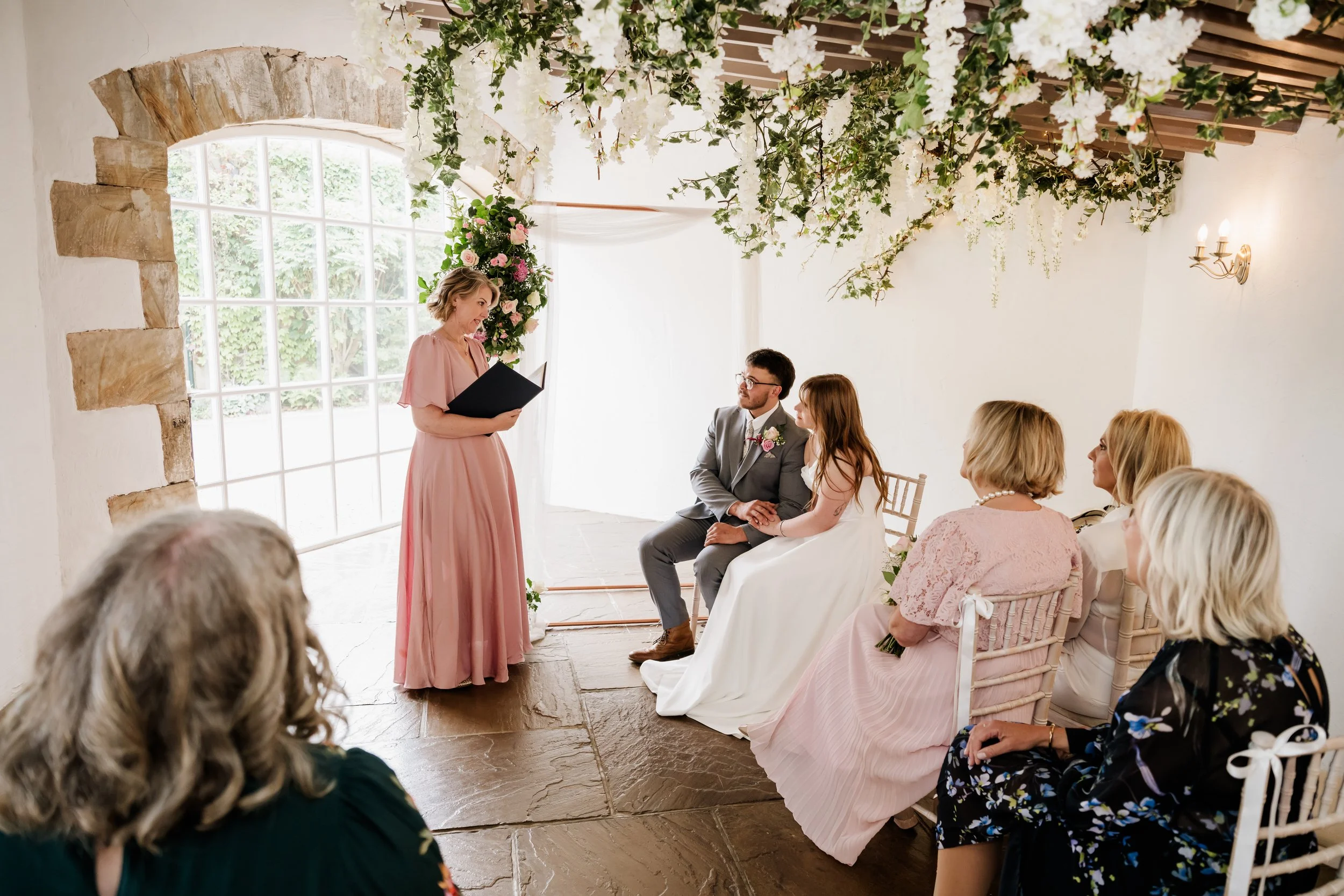 A wedding ceremony taking place indoors near a large window with white framing. The bride and groom are seated.