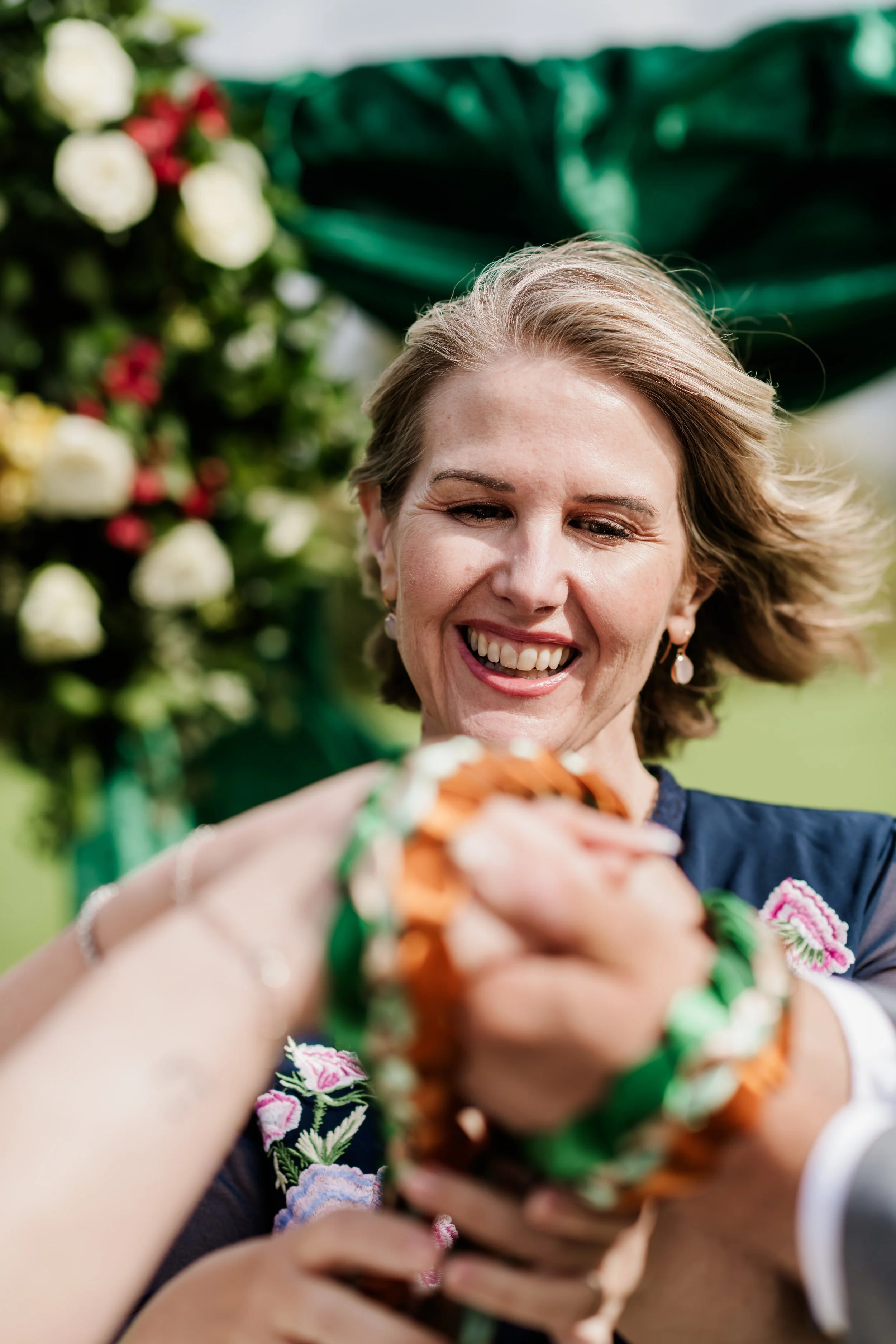 Tammy smiling and binding a couples hands during a handfasting ceremony, with colorful flowers and green leaves in the background.