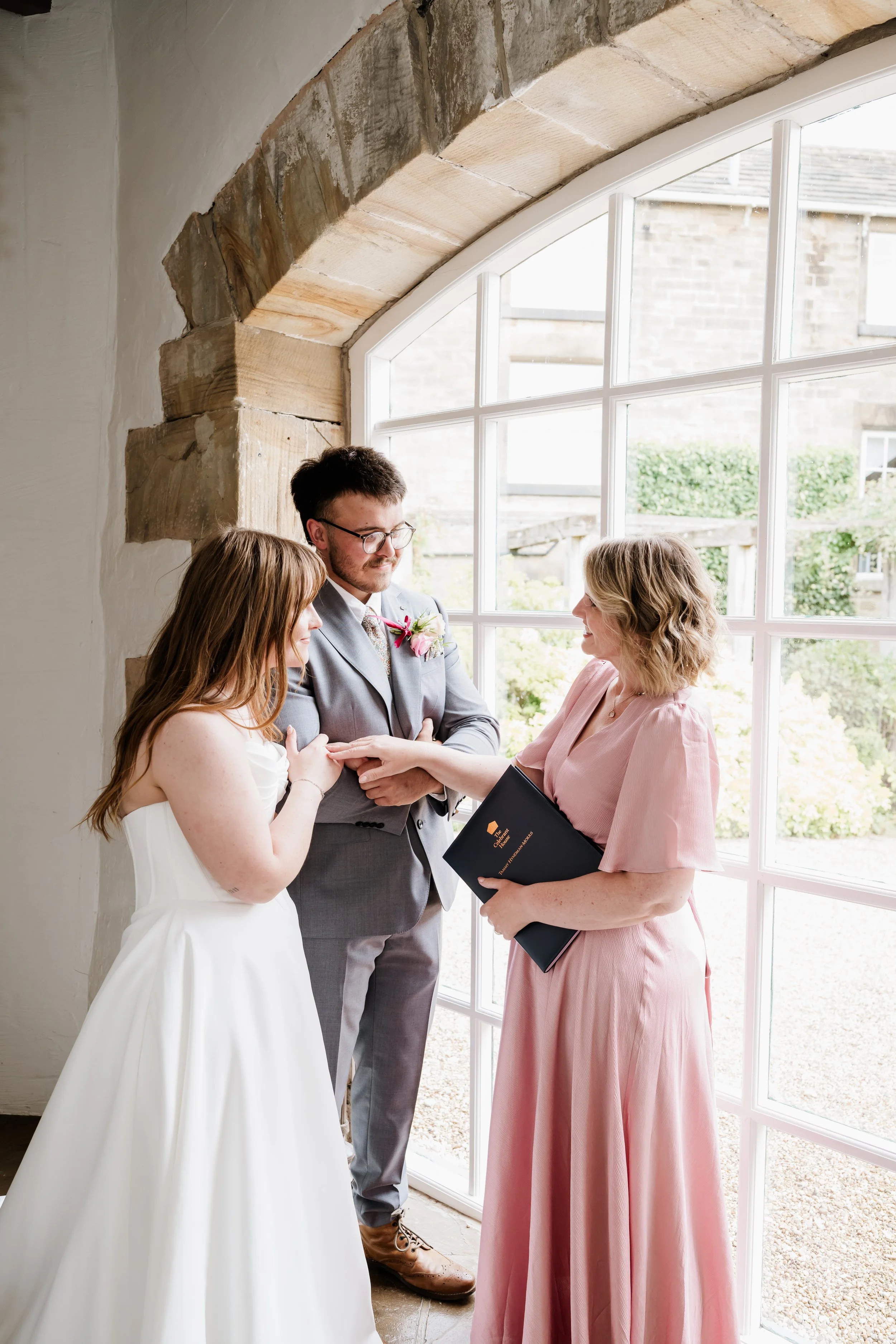 A wedding ceremony where a couple is holding hands, facing Tammy, a female celebrant, next to a large window with a garden view. The bride is in a white dress, and the groom is in a gray suit with glasses.