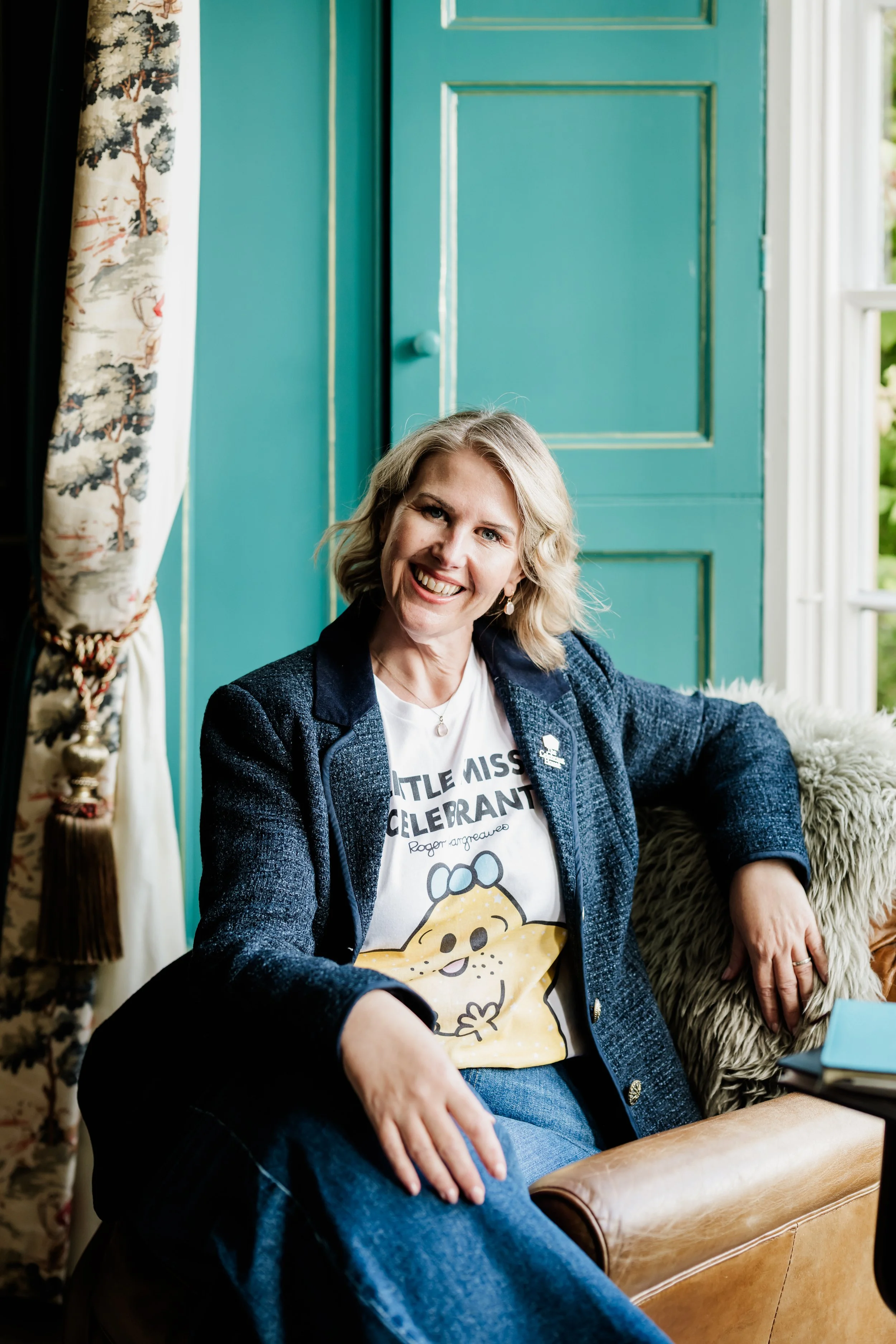 Tammy on her own, she has blonde hair,wearing a graphic t-shirt and dark blazer, sitting on a leather chair inside a room with a teal door and a window with white curtains. She is smiling at the camera.