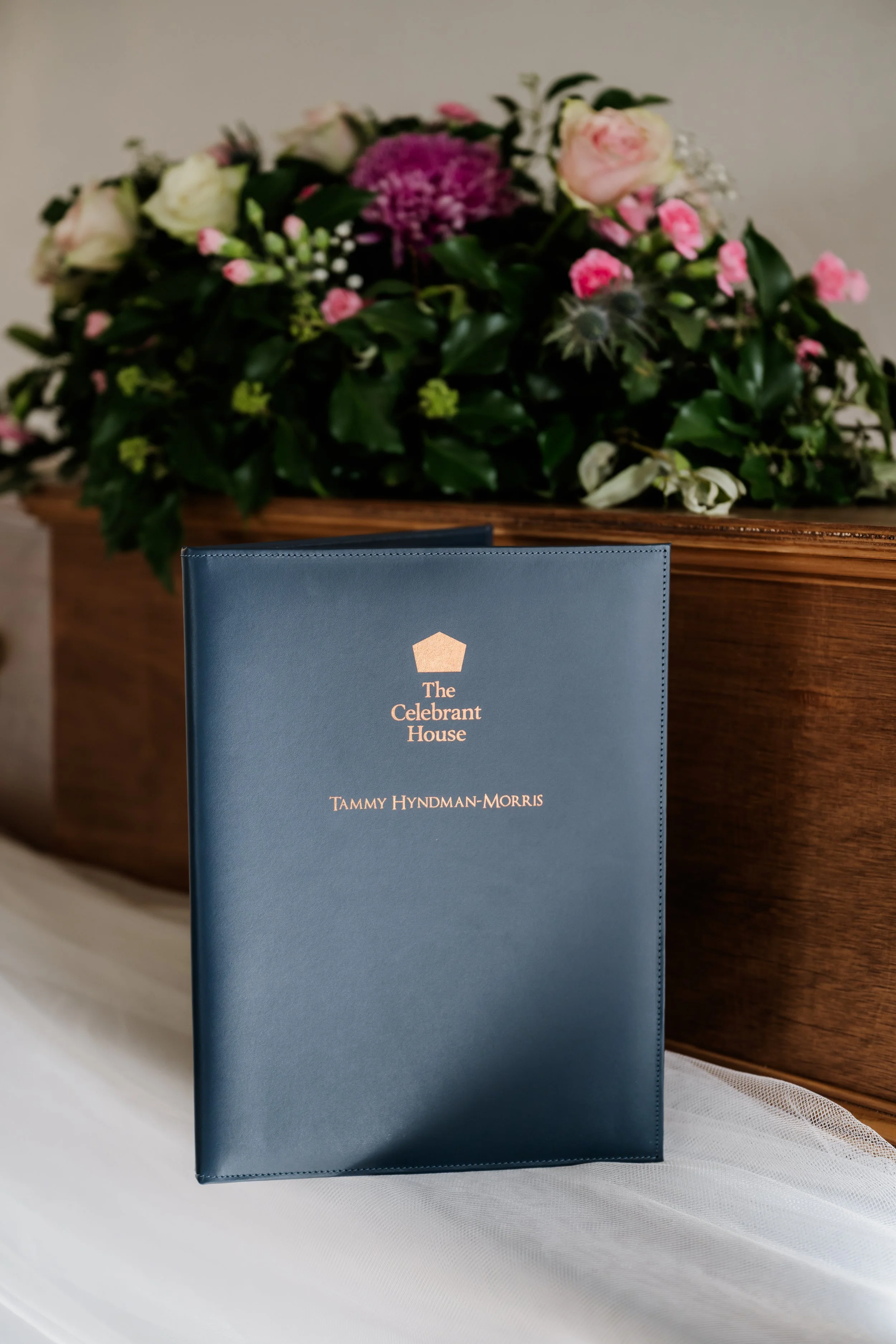 A blue ceremony folder titled "The Celebrant House" is in the foreground. Behind it, there is a floral arrangement with white, pink, and purple flowers on a coffin against a beige wall.