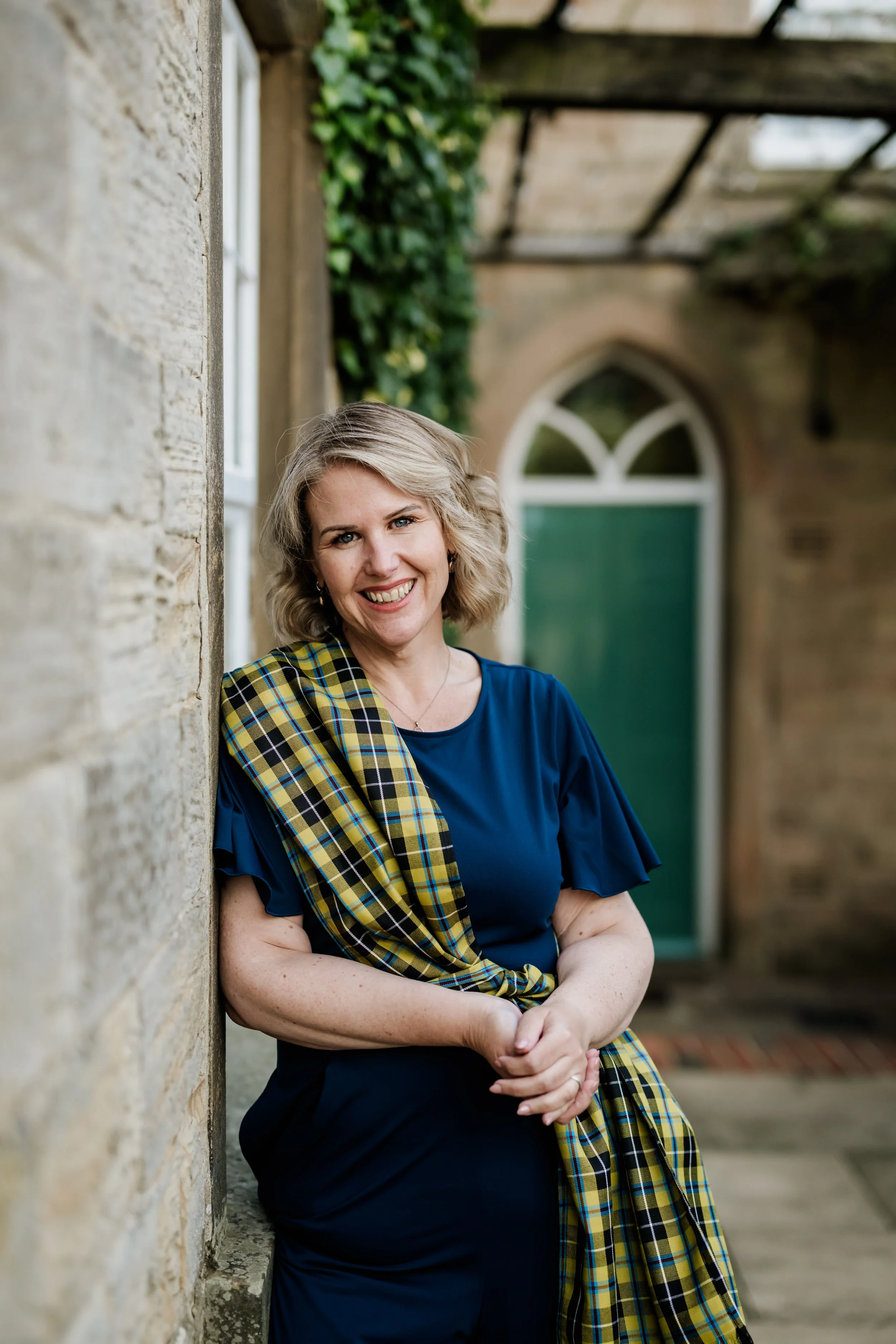 Tammy, a blonde woman with curly hair, smiling, wearing a navy blue clothes and a Cornish Tartan sash, standing outside on a stone house with a brick wall and greenery in the background.