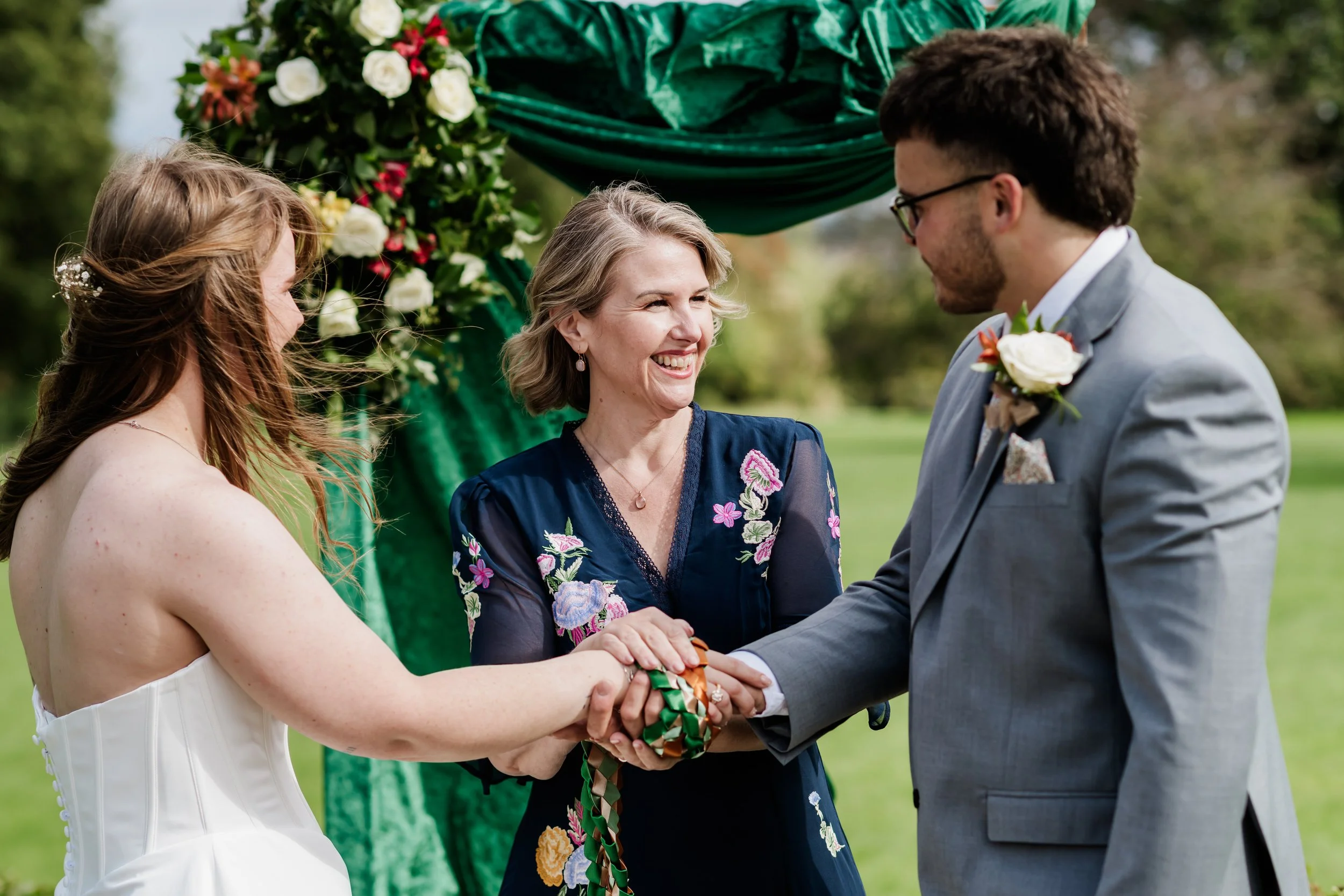 A wedding ceremony outdoors with a bride and groom holding hands, exchanging vows, and smiling at each other, Tammy smiles at them.