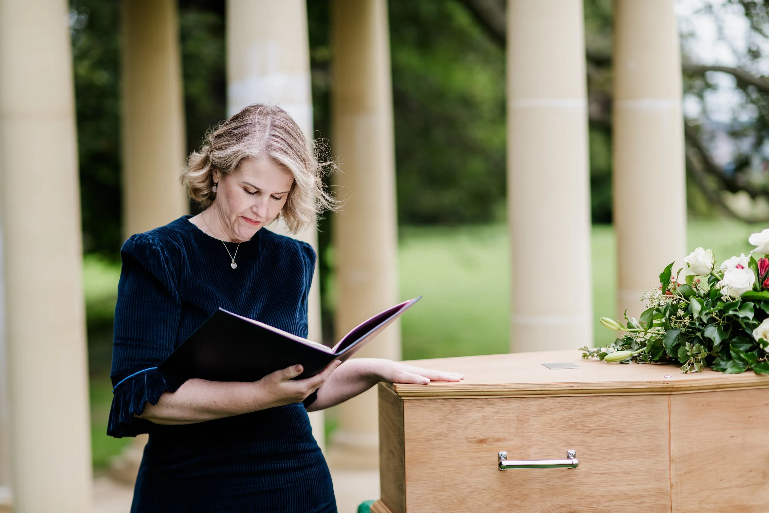 Tammy. a celebrant with blonde hair in a blue dress stands outdoors, reading a book in front of a coffin with a flower arrangement, a foley and green trees in the background.