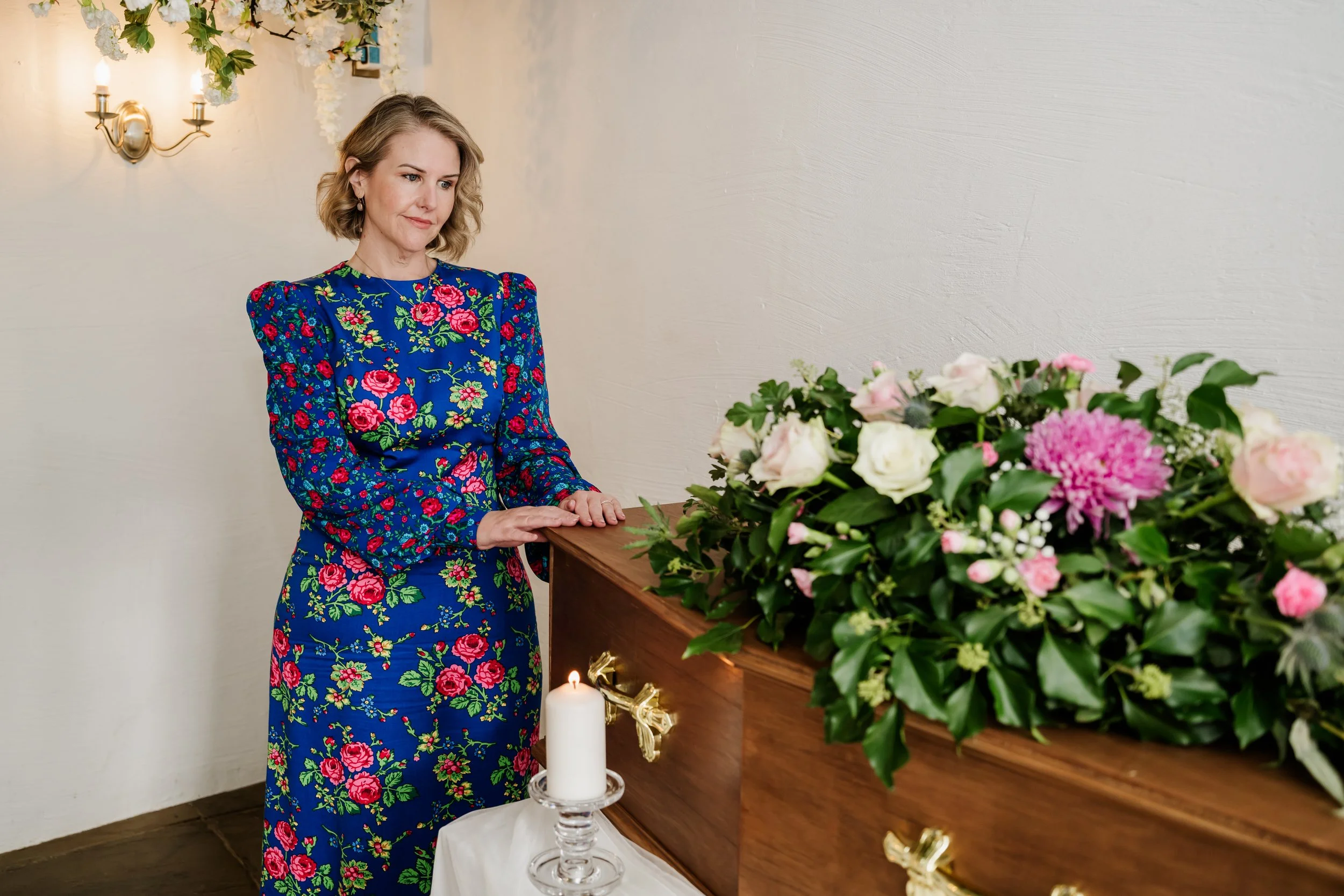 Tammy in a colorful floral dress standing in front of a coffin with flower arrangement and a candle, with a white wall and wall sconce in the background.