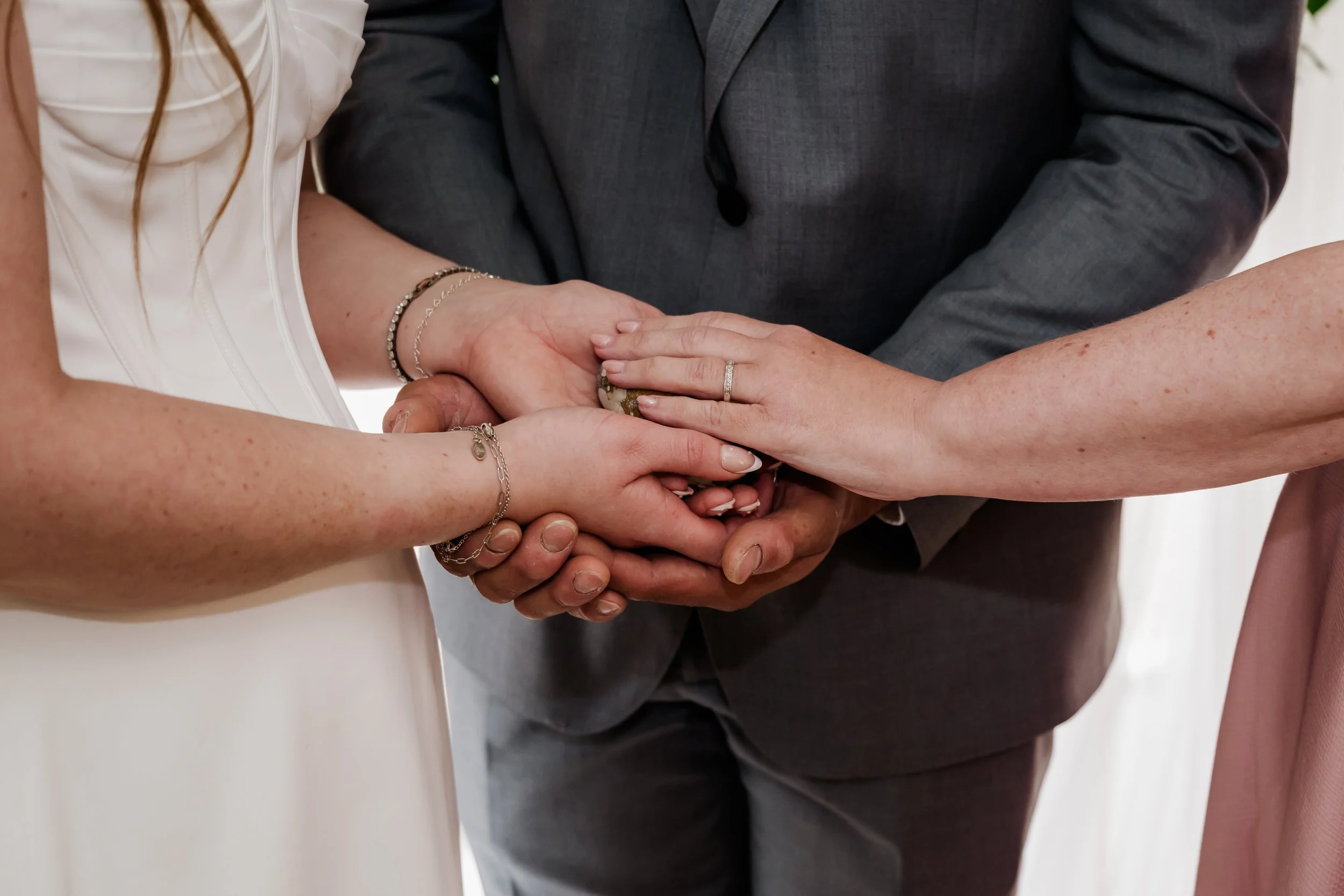 A couple holding hands over an oathing stone with Tammy performing a blessing, with the woman wearing a white dress and jewelry, and a man in a gray suit, at a wedding.