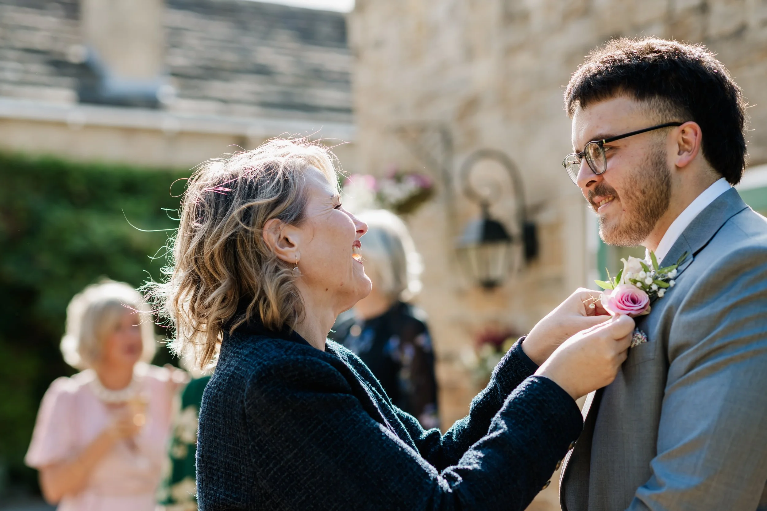 Tammy helps the groom with his buttonhole placement to calm his nerves, smiling at each other at an outdoor wedding ceremony, with a blurred background of other guests and stone buildings.