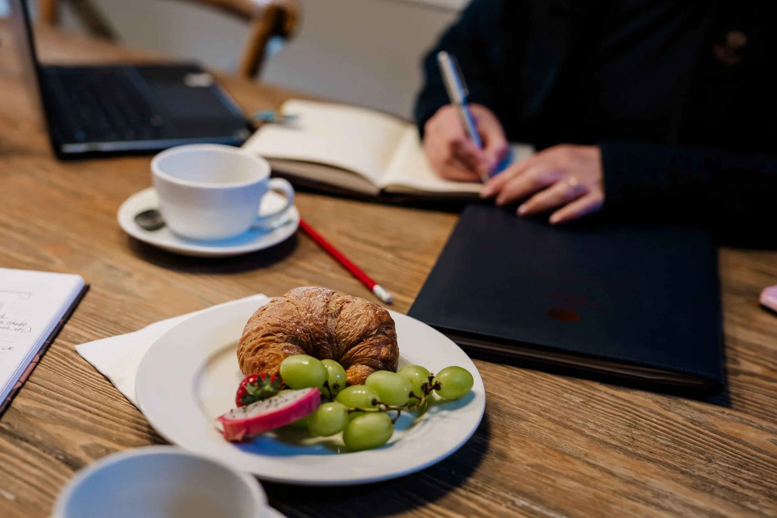 meeting scene on a wooden table with a plate of green grapes, strawberries, and a croissant. There is a cup of coffee, an open notebook, a black laptop, and Tammy is writing in a notebook with a pen.