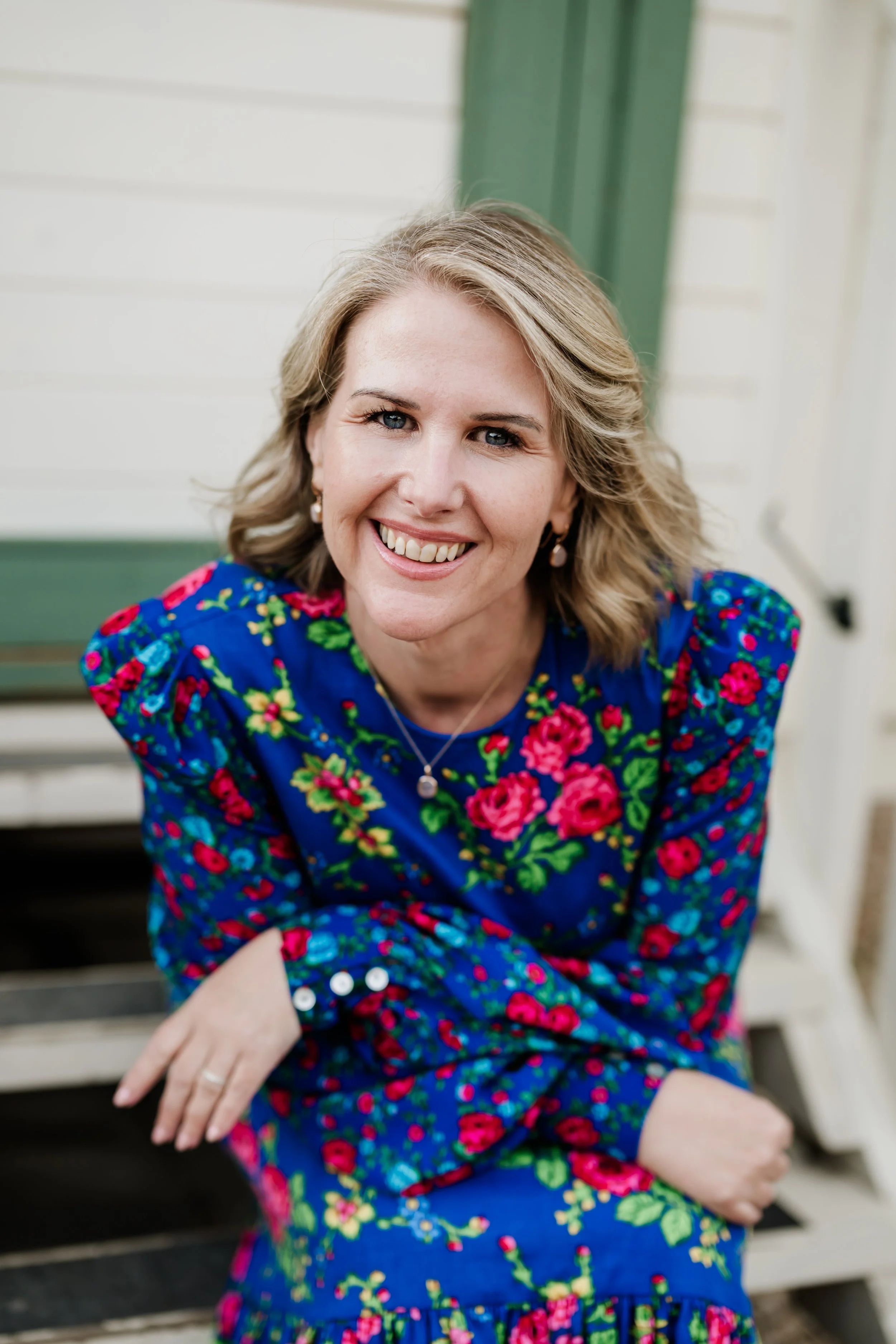 Tammy is smiling, she has blonde hair, blue eyes, wearing pearl earrings and a blue dress with a pink and green floral pattern, sitting on outdoor stairs with a green and white house in the background.