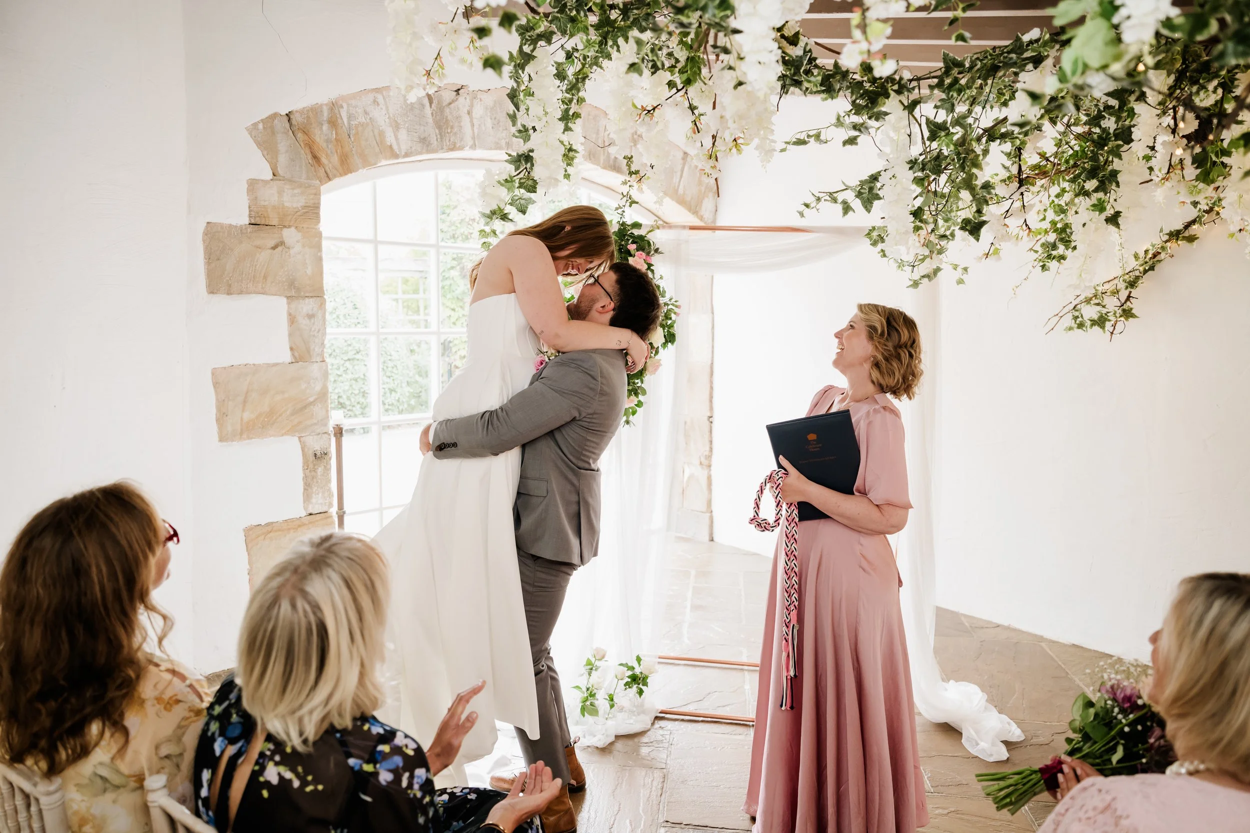 A bride and groom share a kiss during their wedding ceremony, with guests seated and watching, in a decorated indoor venue with floral arrangements and large windows.
