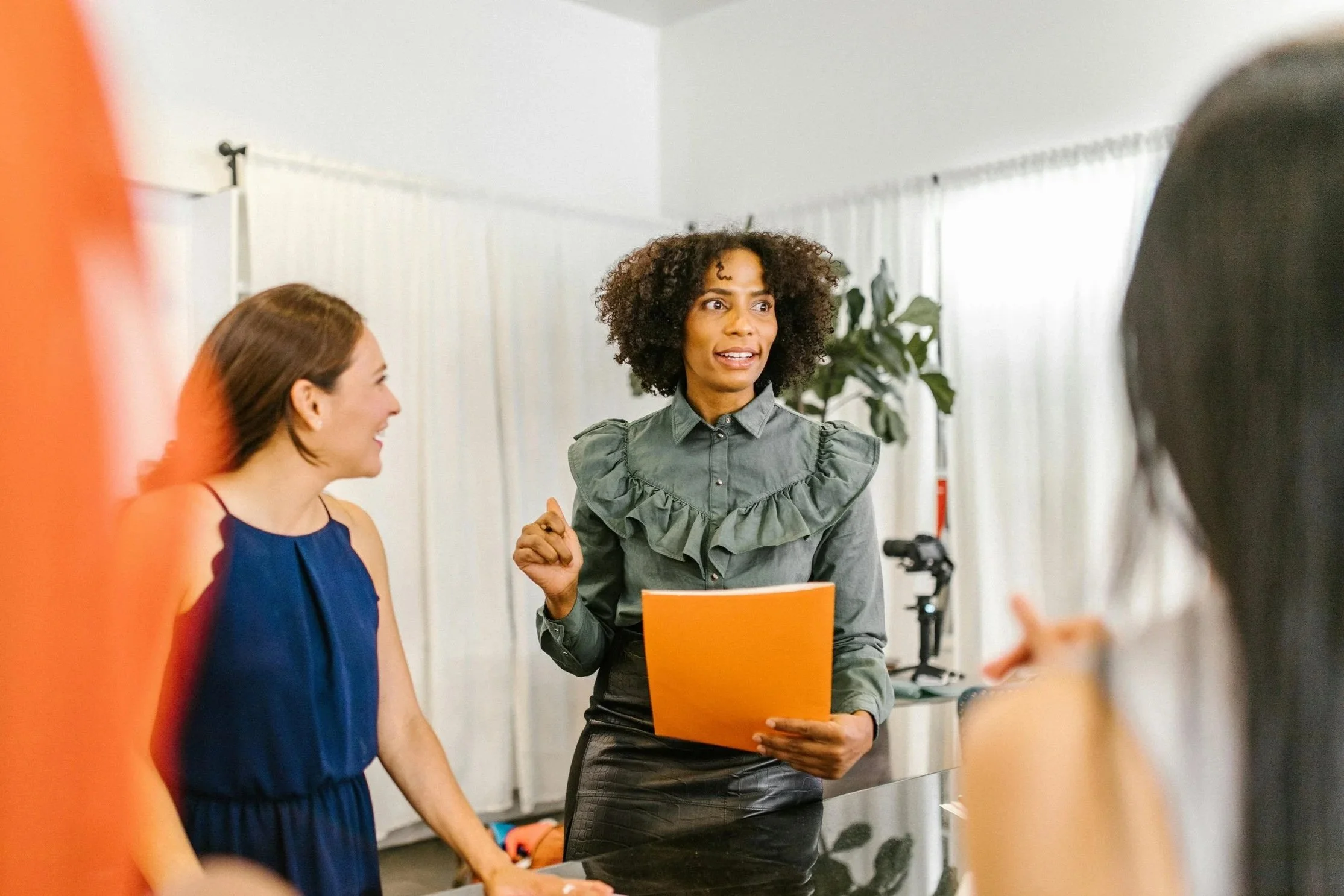 Dames die in een kamer met witte gordijnen en planten een gesprek voeren, onder leiding van een vrouw die een oranje map vasthoudt.