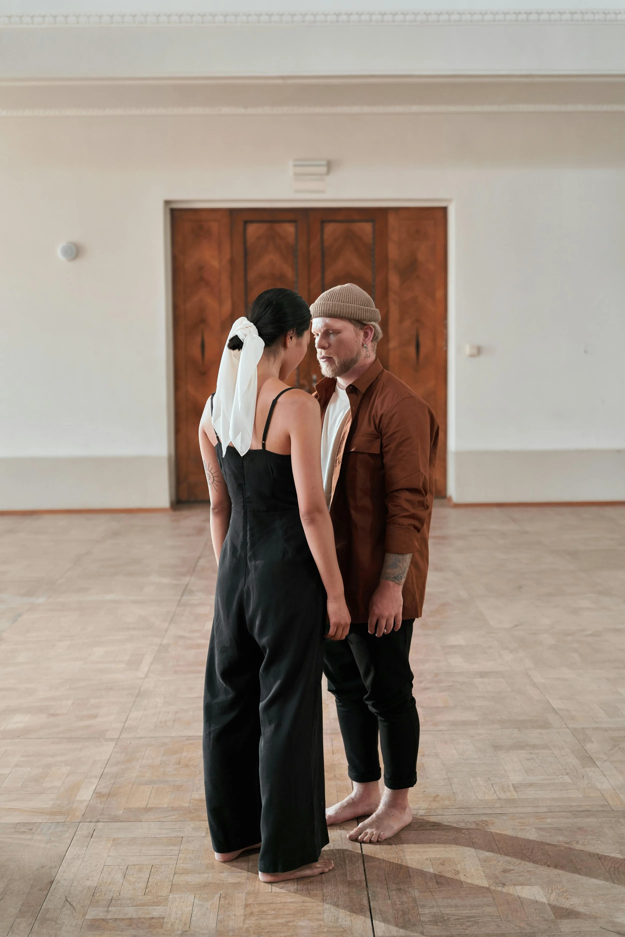Een man en vrouw staan dicht bij elkaar in een kamer met houten vloer en dubbele houten deuren op de achtergrond. Ze kijken elkaar intens aan, en zijn bezig een workshop te volgen om balans in hun relatie te herstellen.