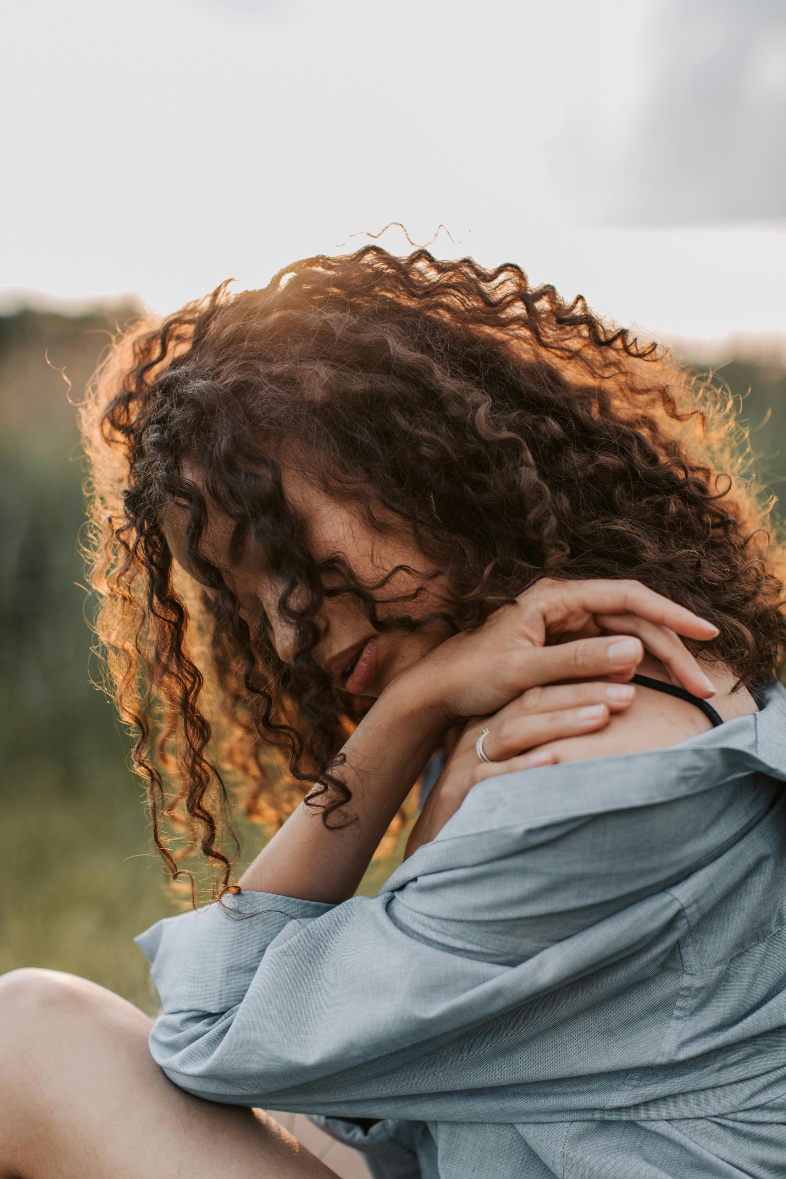 Close-up van een vrouw met krullend bruin haar, zittend buiten met de ogen dicht, hand op haar nek, in een lichte blouse, met een blurred groene achtergrond.