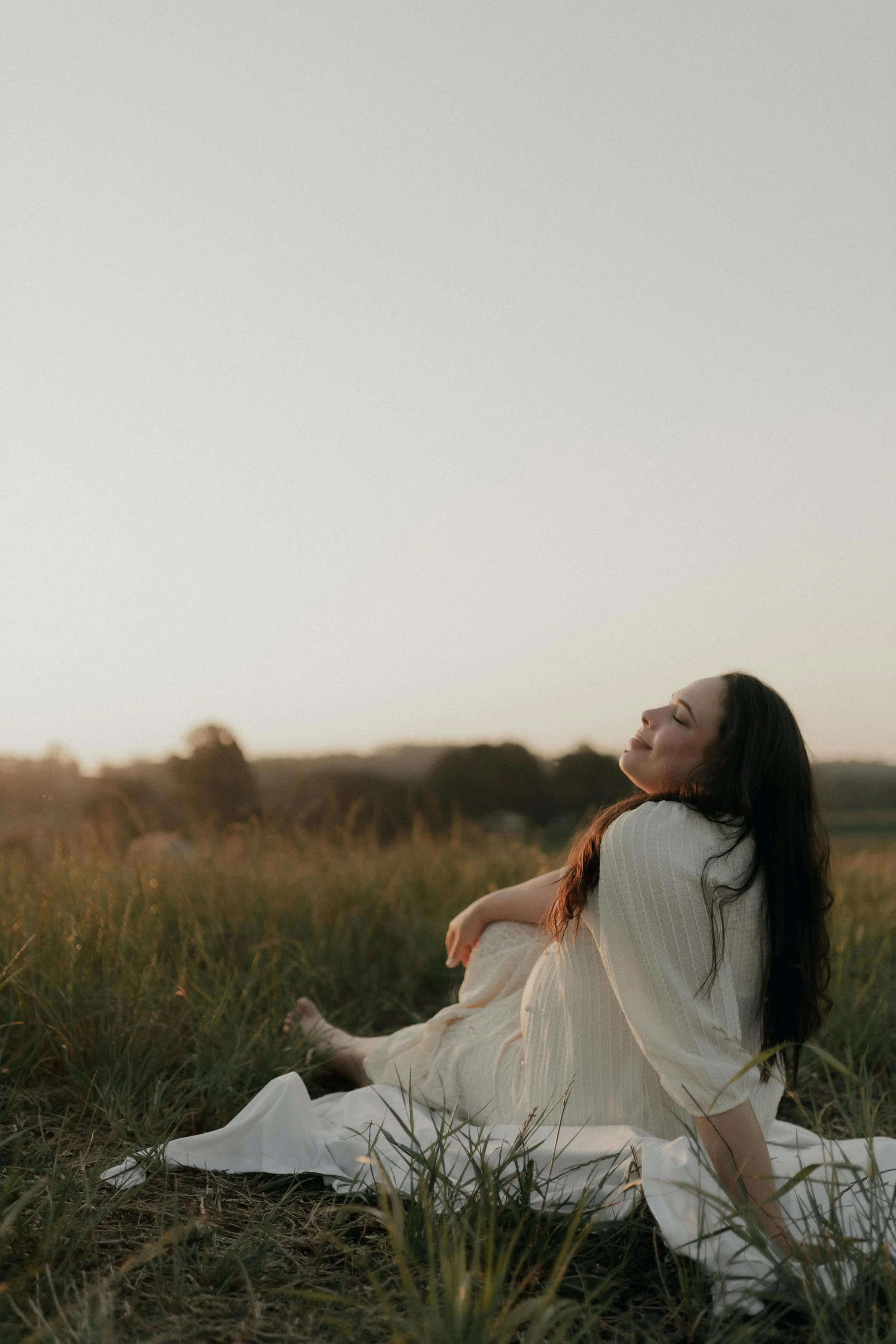 Vrouw zit in een veld met gras tijdens zonsondergang, glimlachend met gesloten ogen. Ze heeft een rustige, serene uitstraling.