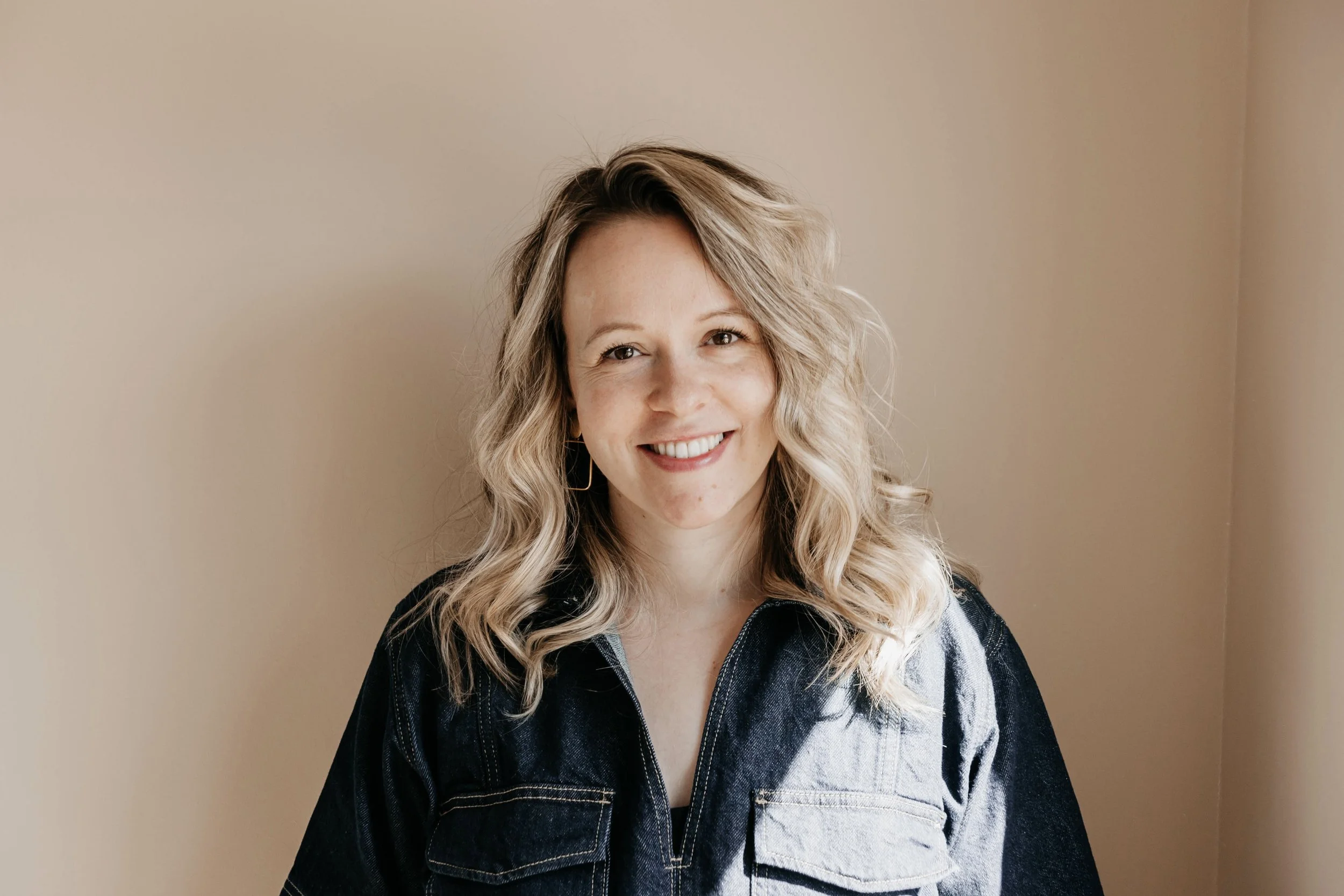 Portret van een lachende vrouw met blonde, gekrulde haar, gekleed in een donkerblauwe en lichte spijkerjack, staand voor een beige muur.
