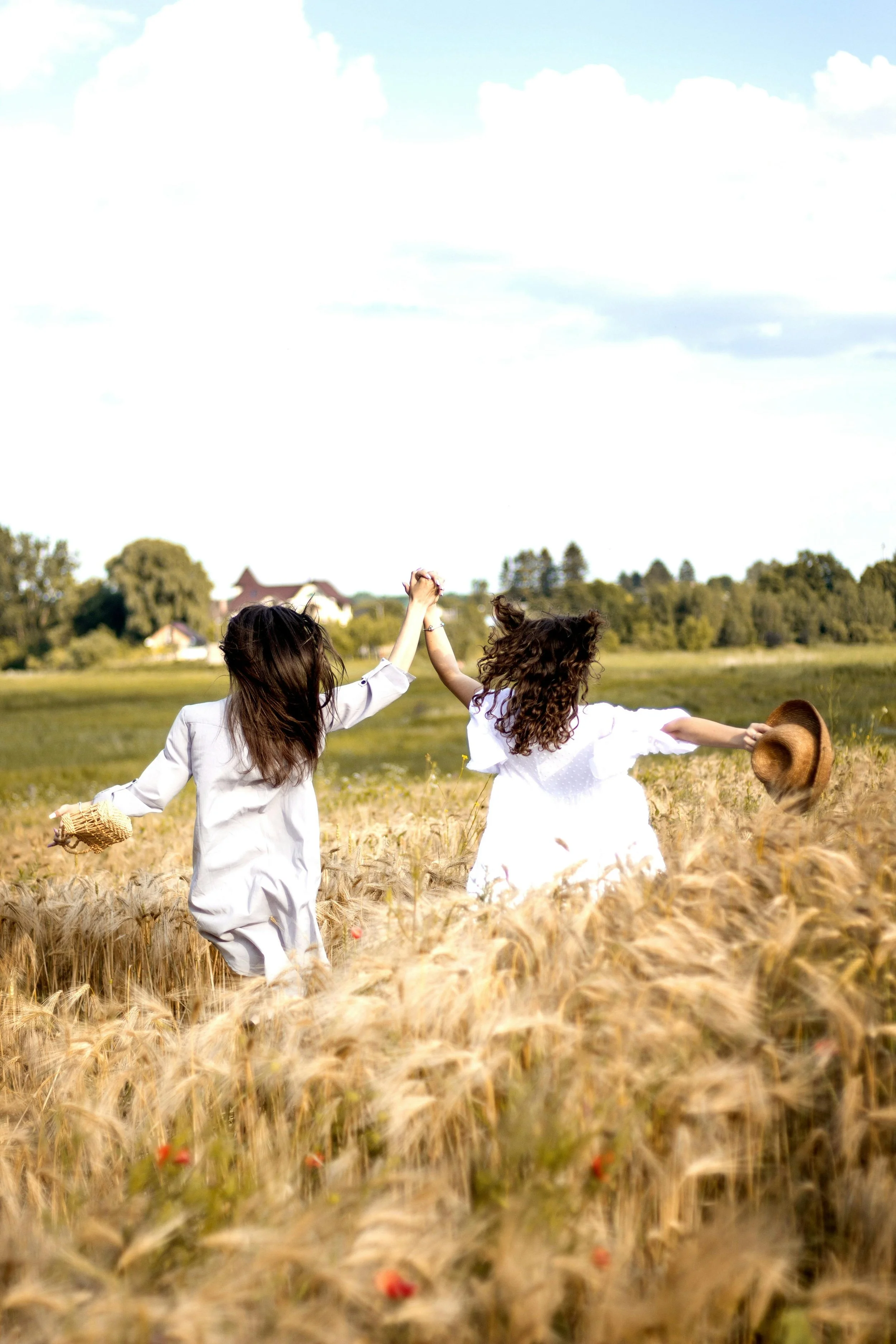 Twee vrouwen dragen witte jurken en rennen tarweveld, terwijl ze elkaar vasthouden en lachen, met groene bomen en huizen op de achtergrond onder een blauwe hemel met wolken.